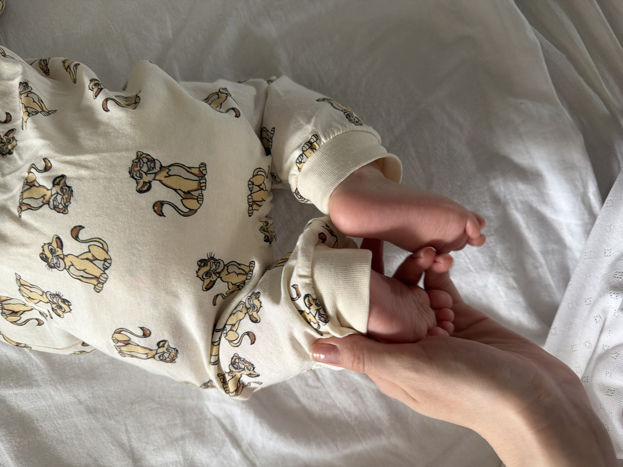 A close-up photograph captures a baby lying on a white fabric surface, likely a bed. The baby is wearing a cream-colored onesie adorned with a repeating pattern of Simba from "The Lion King." The focus is on the baby's bare feet, which are being gently held by an adult hand. The hand has a light skin tone and a glossy manicured fingernail. The background consists of the crumpled white fabric. No location data is detectable.