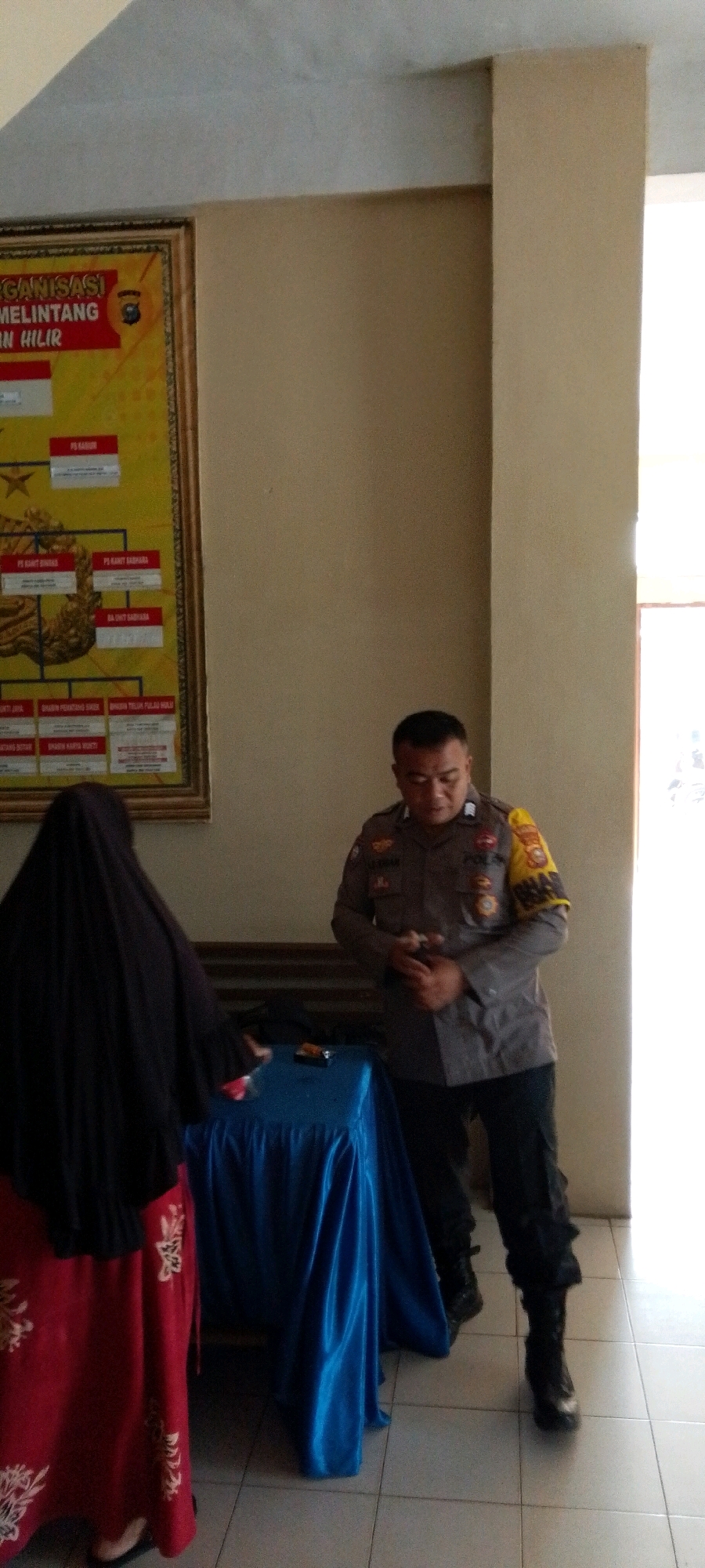 A uniformed male police officer stands in front of a table draped with a blue satin cloth, looking down at his hands. To his left, a person wearing a black hijab and a red and white patterned skirt is partially visible, facing away from the camera and towards the table. Behind them, an organizational chart hangs on the wall. The floor is tiled. The scene appears to be indoors, possibly in an office or a community center.