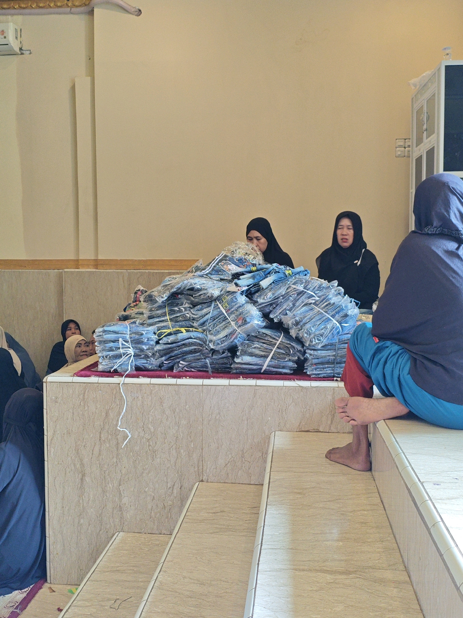 In Teddaopu, Indonesia, a group of people are gathered in a room with light-colored walls and a patterned tiled floor. In the foreground, a person wearing a dark blue headscarf and red and blue pants sits on a tiled step, facing away from the camera. To their left, a large pile of clothing items, individually wrapped in plastic and tied with white string, is stacked on a raised platform. Several individuals are partially visible behind the pile of clothing, some wearing headscarves. One woman in a black headscarf is looking towards the camera, while another woman in a lighter colored headscarf appears to be speaking. An air conditioning unit is mounted on the wall in the upper left corner of the image. The environment appears to be indoors, possibly a community hall or a place of worship.