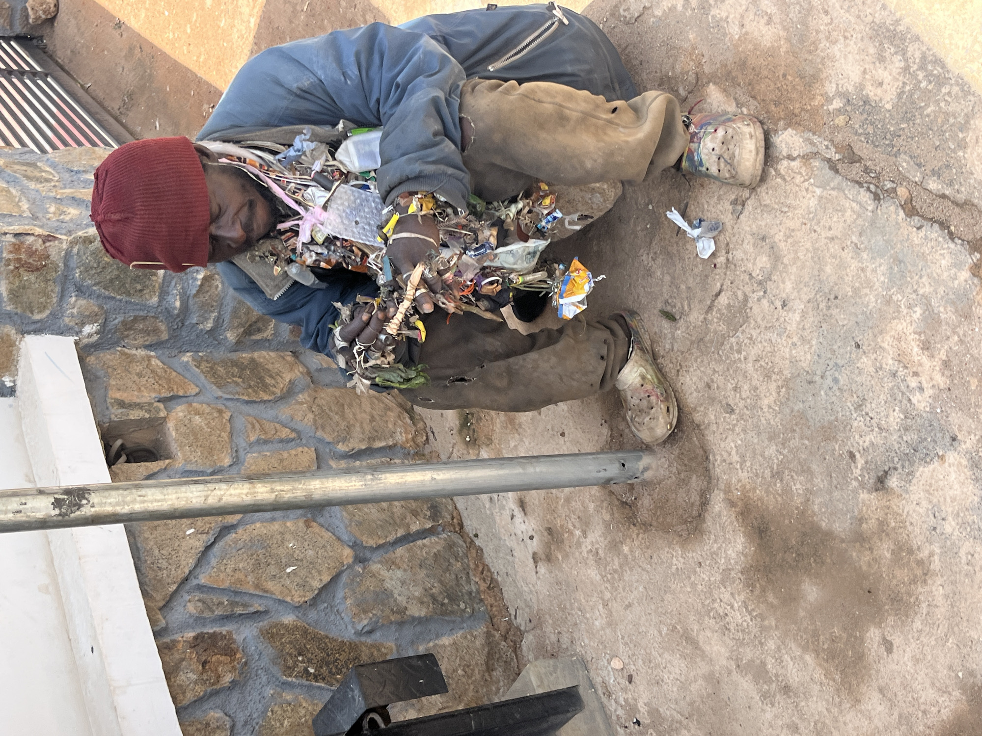 An adult male wearing a maroon beanie, a blue jacket, and brown pants is sitting on a concrete surface next to a stone wall. He has an assortment of items hanging around his neck, including plastic wrappers, small bottles, and what appears to be dried plant material. His right leg is exposed, revealing a hole in his pants. He is wearing a light-colored shoe with colorful accents on his right foot and a similar shoe on his left foot. A metal pole is visible in the foreground, and the background shows a section of a building with a white ledge and a metal fence. The ground is a mixture of concrete and dirt, with some damp patches.