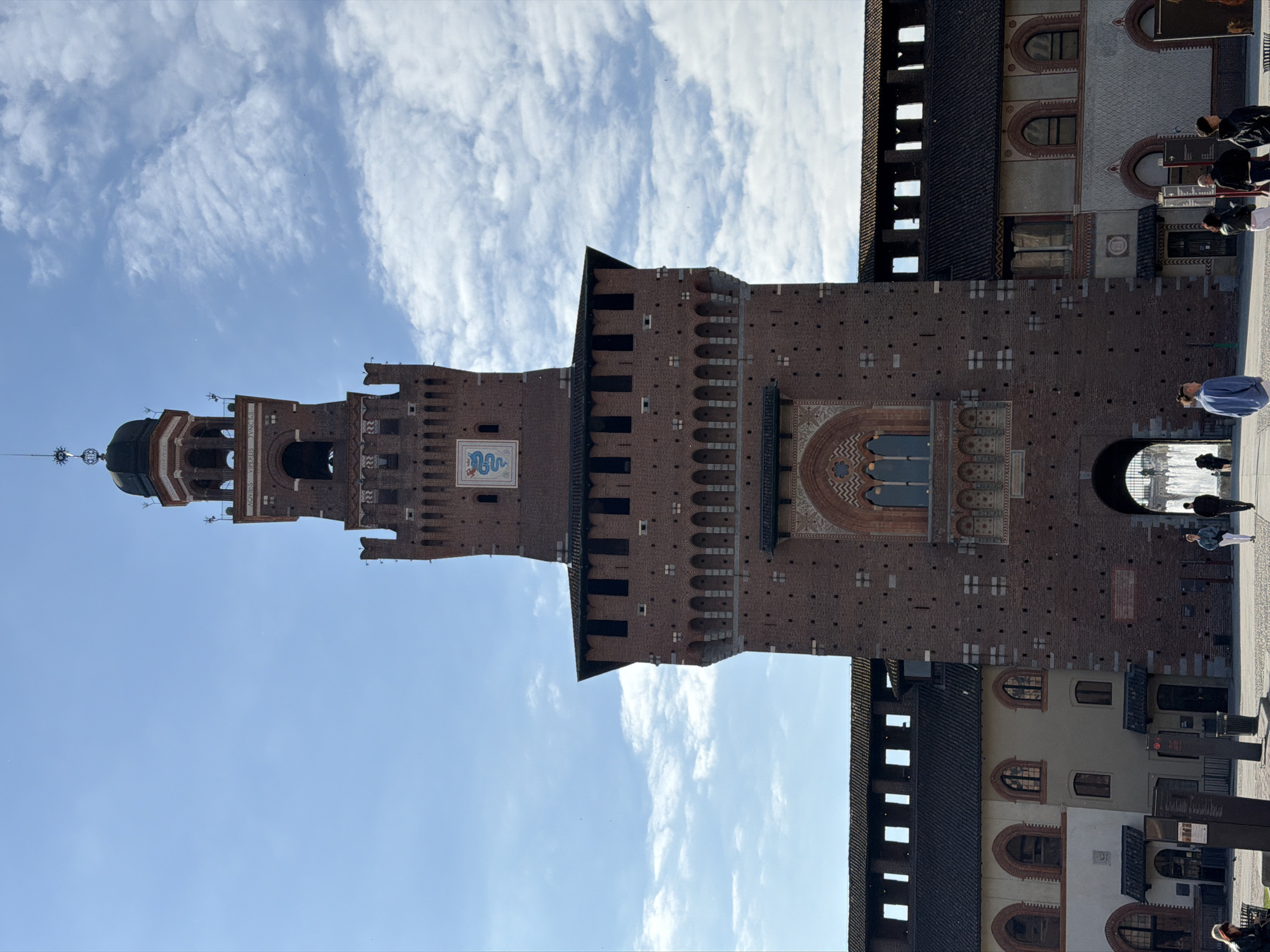 The image depicts the Sforza Castle (Castello Sforzesco) in Milan, Italy. The castle's tower stands prominently against a bright blue sky with wispy clouds. The brick facade and architectural details of the castle are clearly visible. Several people are present in the foreground near the castle entrance. The ground appears to be paved, and there are some signposts visible.