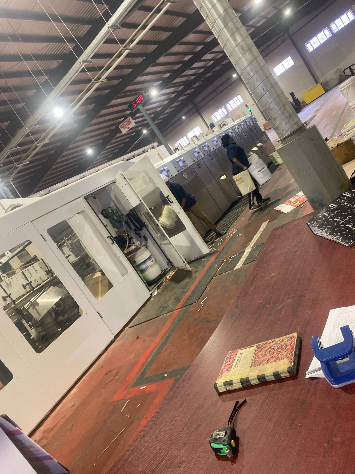 The image depicts an indoor industrial environment, likely a printing or manufacturing facility, situated in the city of Abuja, Nigeria. In the foreground, a dark red wooden table occupies the lower right portion of the frame, displaying a closed patterned book, a blue hole punch, a document, and a small green and black tape measure. The midground is dominated by a large, white industrial machine with glass-fronted sections revealing internal mechanical components. Two individuals are visible near this machine; one is partially obscured behind the machine, and another person in dark clothing is walking away, carrying two white buckets. The floor is a reddish-brown concrete with visible black and red painted lines, possibly indicating pathways or safety zones. Scattered debris, appearing as small white pieces and thin sticks, is present on the floor. In the background, a row of identical grey industrial units with control panels is visible, and a large, ribbed metallic ventilation pipe extends upwards. A digital display showing "5:00" and a sign pointing downwards with the text "PRINTING / FINISHING SECTION" are also discernible. The ceiling is a dark, corrugated metal structure with exposed beams and numerous hanging lights. The overall lighting suggests it is operational during daylight hours.