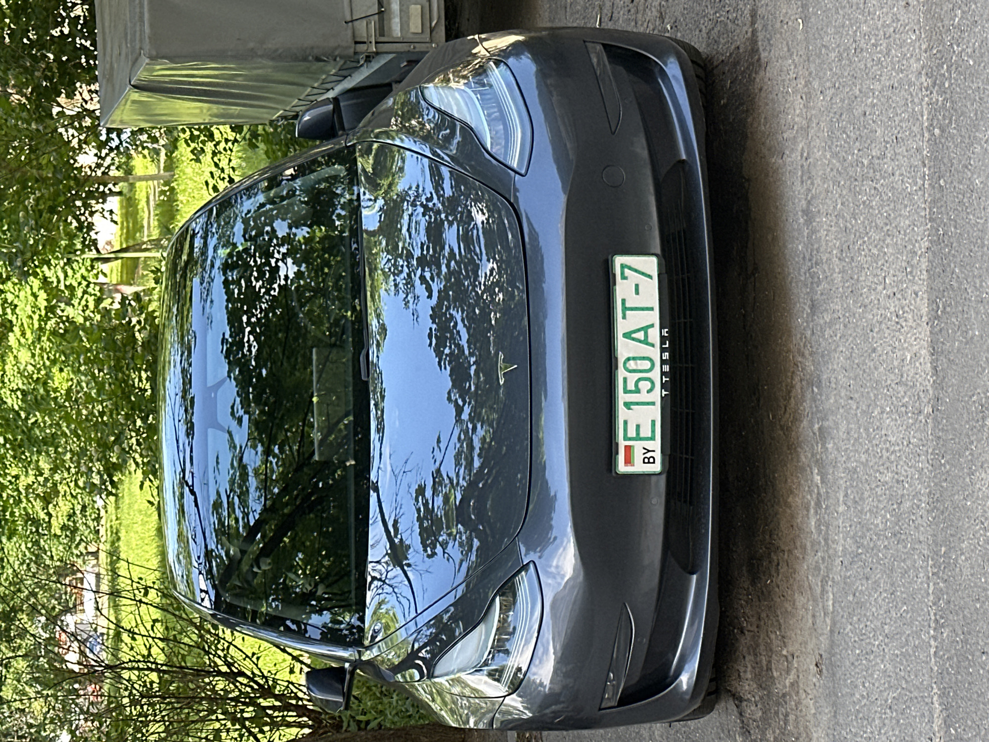 A gray Tesla Model 3 is parked on a paved surface, likely a street or parking lot, in front of lush green trees and foliage. The car's front license plate is visible, displaying "BY E 150 AT-7" and the Tesla logo. The background suggests a residential or park-like setting within a city, possibly Minsk, Belarus, given the "BY" on the license plate. The car is clean and appears to be in good condition. There are no people or other vehicles immediately visible in the frame. The lighting indicates it is daytime, with sunlight filtering through the trees and reflecting off the car's surfaces. To the right of the car, a portion of a gray trailer with a tarp cover is visible.
