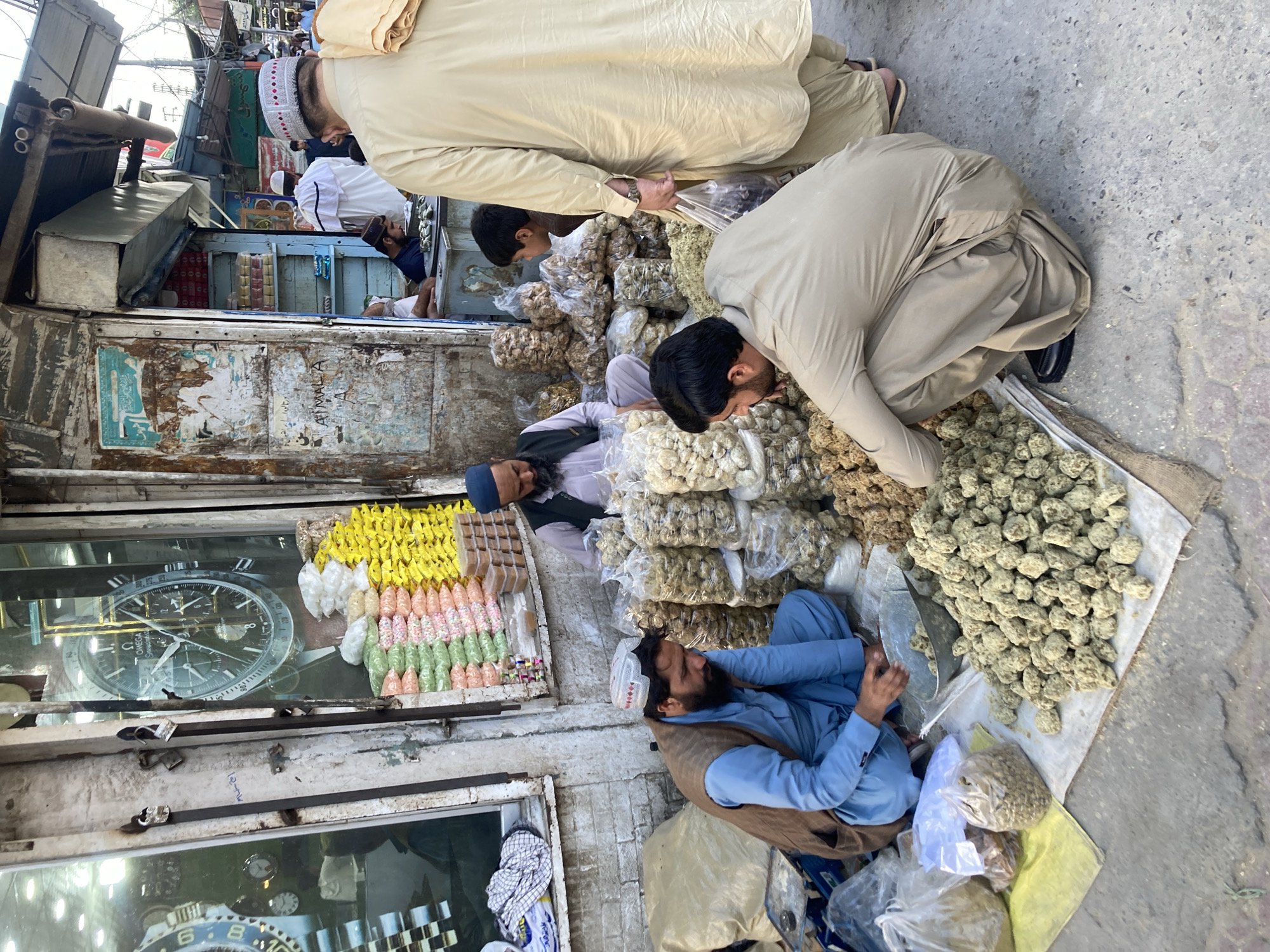 In Quetta City Tehsil, Pakistan, people are gathered at a market stall. The scene depicts a vendor selling dry fruits. The stall is stocked with an array of colorful candies, nuts, and dried fruits. One man is kneeling down next to the pile of goods, while another is kneeling near the merchandise. The stall is in a rundown area of the city.