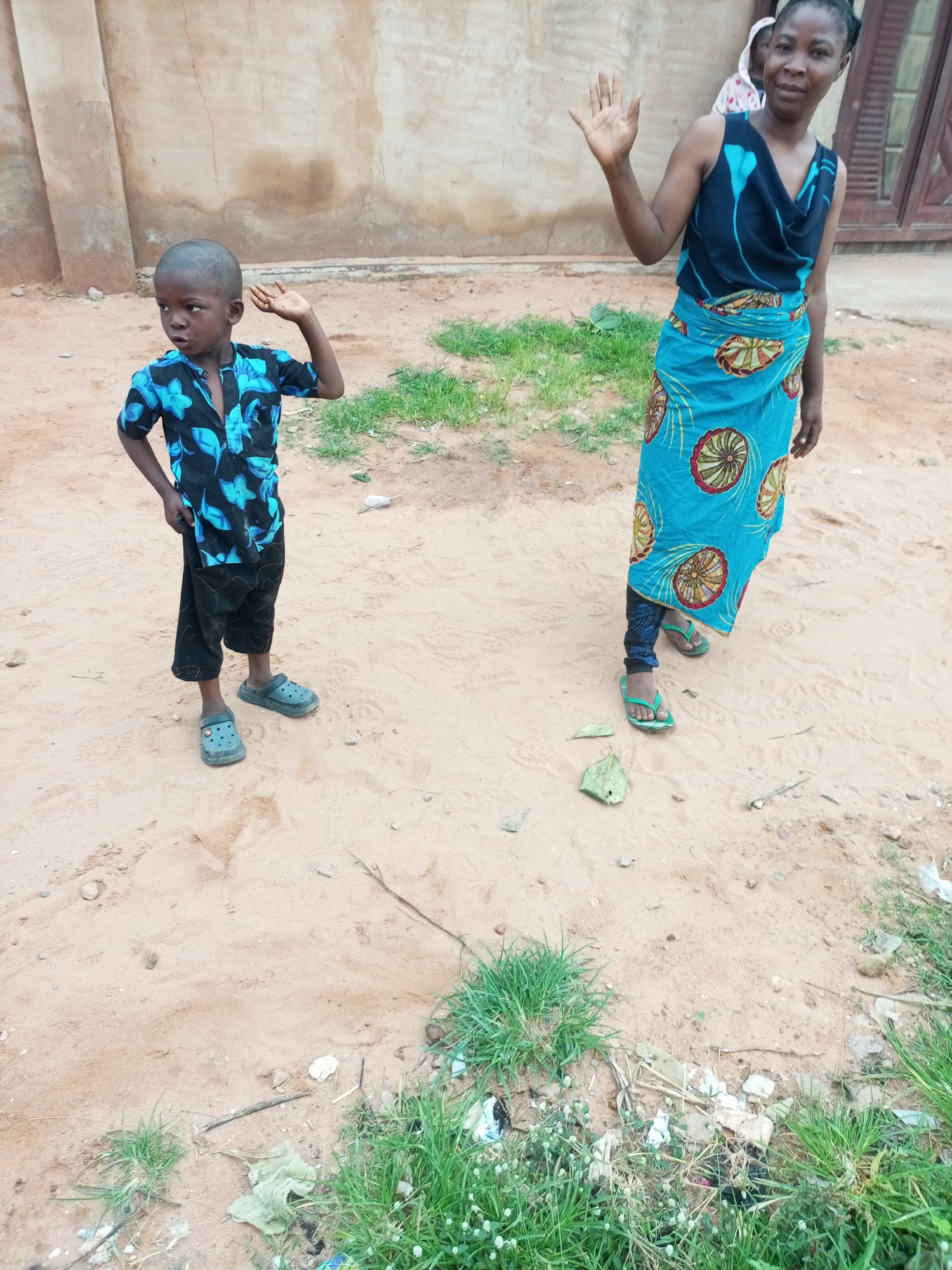 In Tempure, Nigeria, a young boy stands on a dirt ground, wearing a blue patterned shirt and black shorts. His left hand is on his hip, and he looks to his left. To his right, a woman waves with her right hand, looking at the camera. She is wearing a dark blue dress with a floral pattern and a vibrant blue wrap skirt adorned with circular designs. Behind her, a person is partially visible, wearing a light-colored hooded garment. The background features a beige, textured wall, and the ground is covered with sand and patches of green grass. Debris, including paper and plastic, is scattered on the ground near the grass.