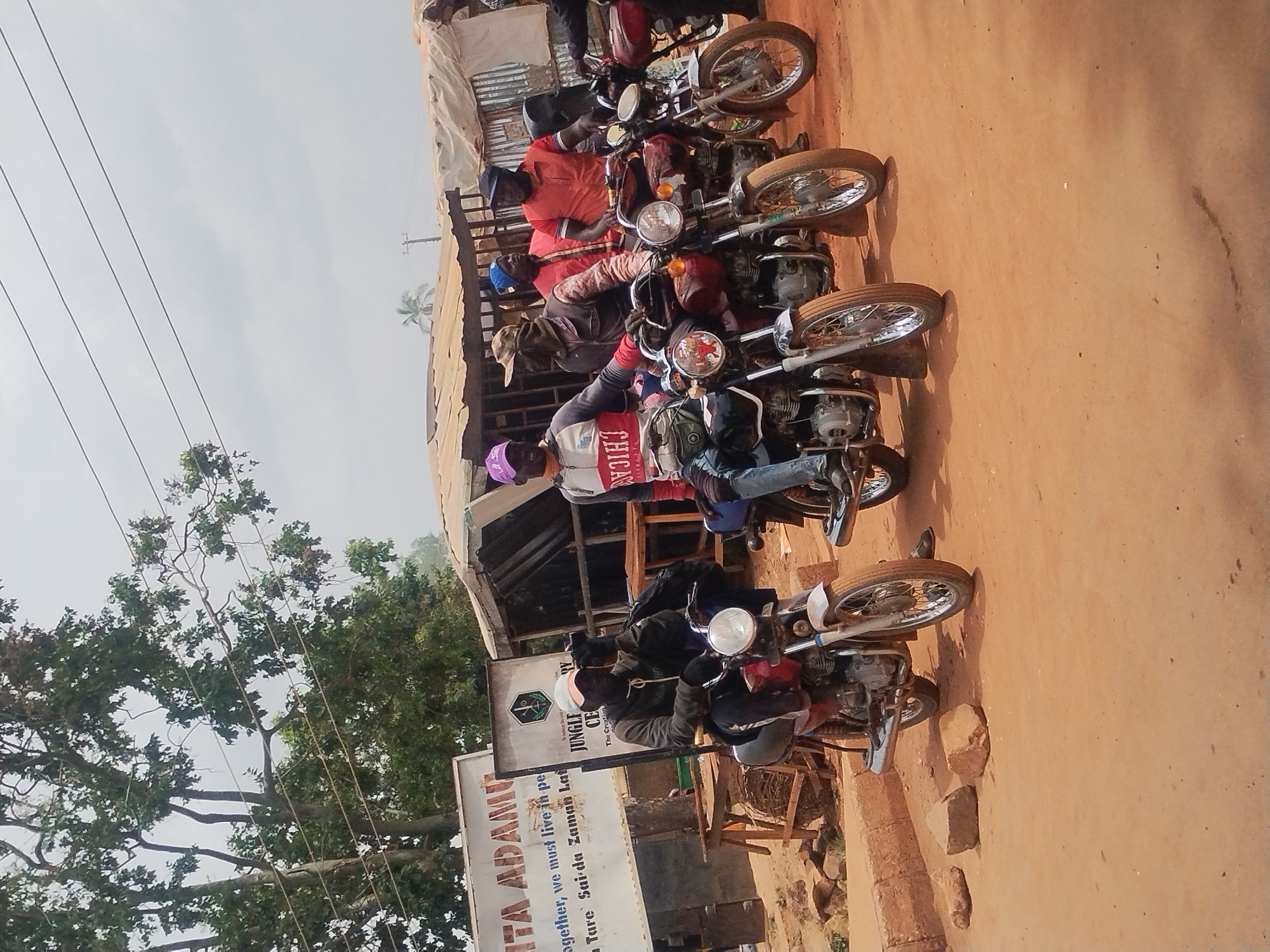 The image shows five individuals seated on motorcycles parked along a dirt road next to a building, likely a shop or business, in a tropical location. The building has a sign with the text "ITA ADAMO, Together, we must live in pe Tare Saida Zaman Lat" and another sign that reads "JUNGLE CE, The Creation, BY AVJIHI." The riders are casually dressed; one wears a purple cap and a "Chicago" branded jacket, while others sport hats and various colored shirts. The motorcycles are positioned perpendicular to the road, on what appears to be a dirt surface. A large tree with dense foliage is visible to the left of the building. Considering the signs and visual cues, the location is plausibly within Jos, Nigeria.