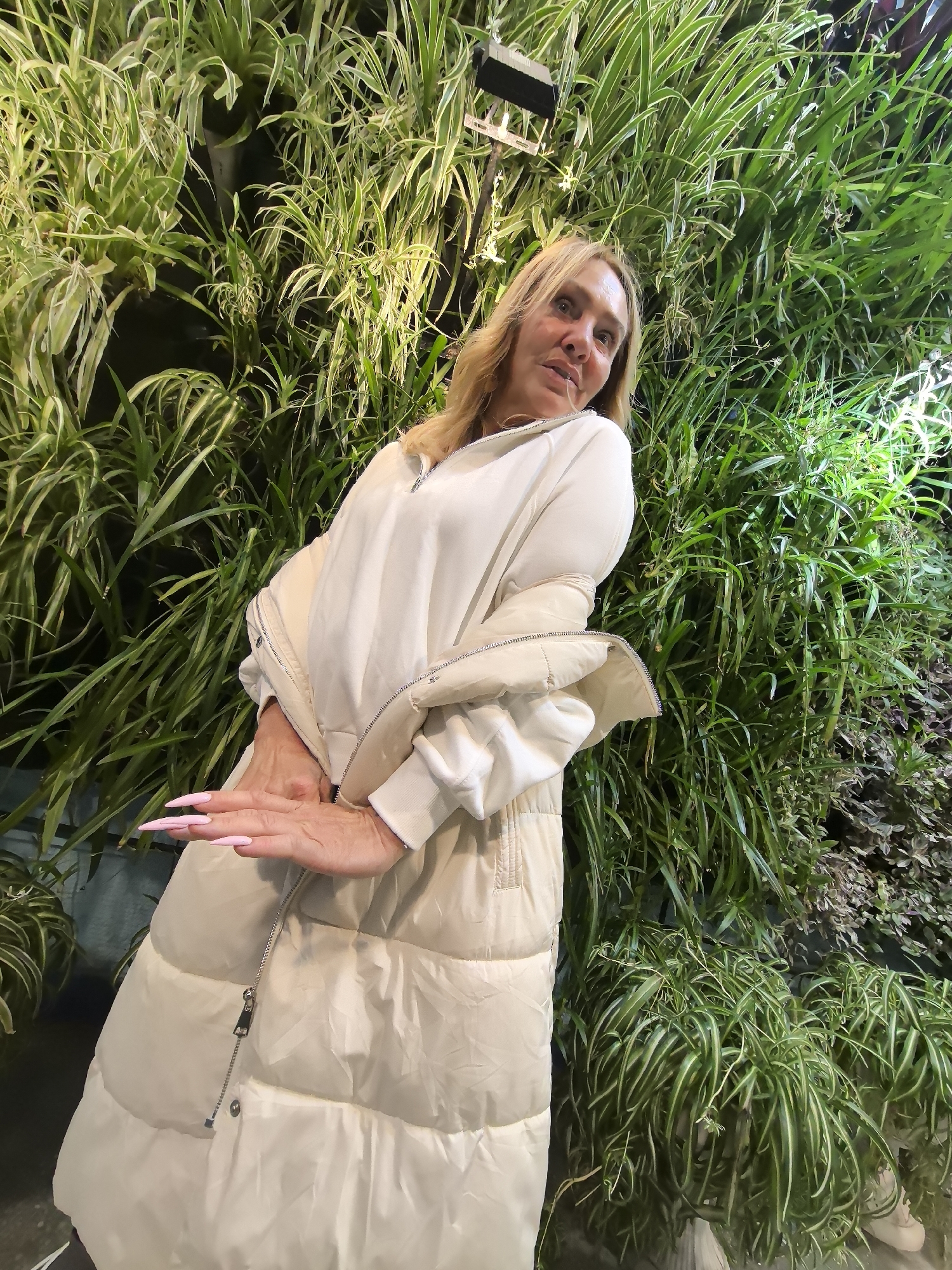 A woman stands in front of a green wall with plants. She has long, pink nails and is wearing a light-colored puffer jacket over a white sweatshirt. The green wall is composed of different varieties of plants with long, thin leaves. There is a light fixture at the top of the green wall. The setting could possibly be Izhevsk, Russia.