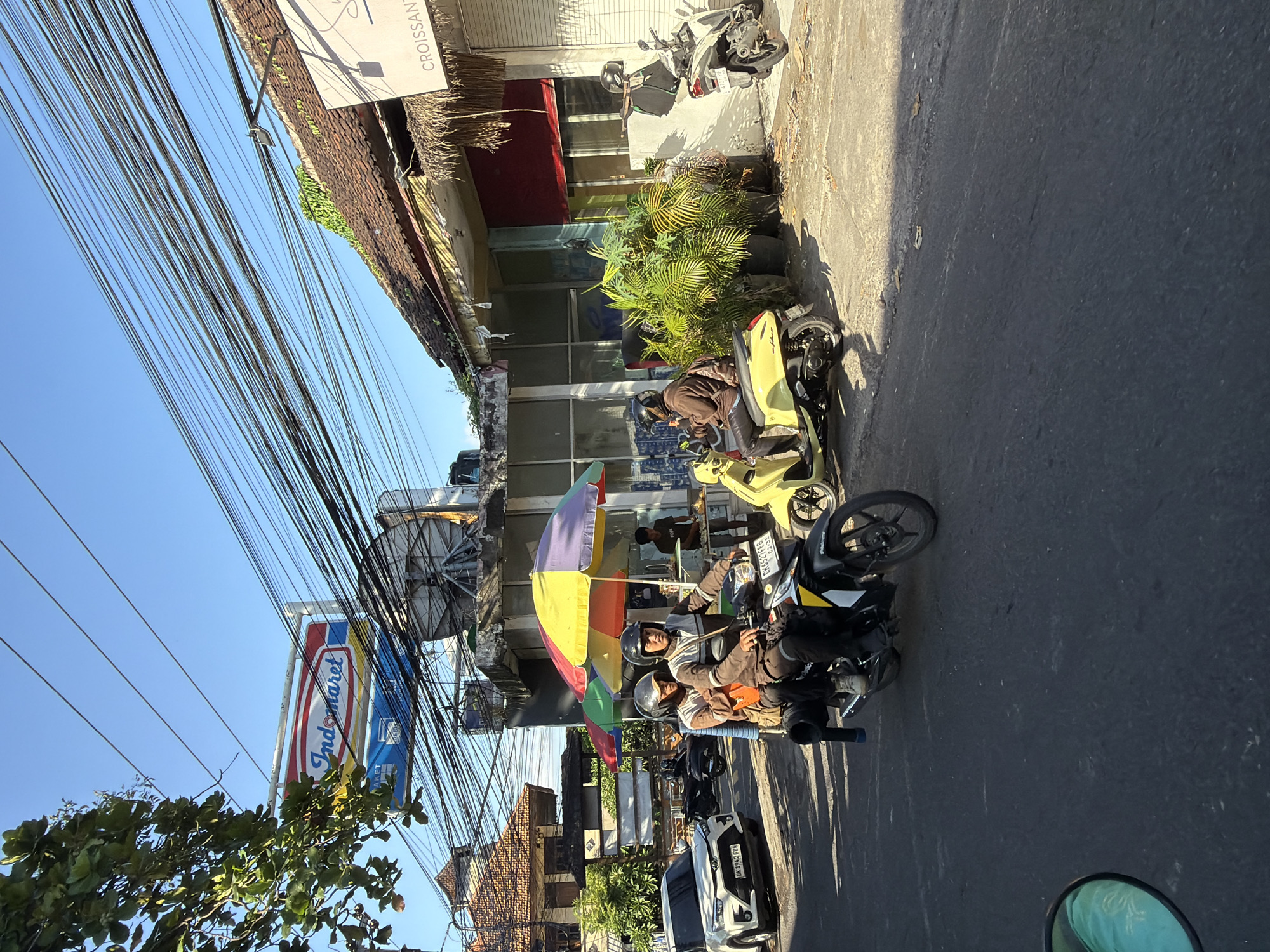 The image captures a street scene in North Kuta, Indonesia, on a clear, sunny day. The foreground features a dark asphalt road. A black motorcycle with two occupants, both wearing helmets and casual clothing, is the primary subject. The rider in front wears a brown and white striped jacket, and the passenger behind wears a brown jacket with an orange bag slung over their shoulder. The motorcycle has a license plate that reads "DK 6521 FBH". To the right of this motorcycle, a yellow scooter is parked, with a person in a brown jacket and helmet seated on it, facing away from the camera.
Further down the street, a white SUV is parked on the left side of the road, its license plate reads "DK 1962 FBH". Behind the motorcycles and scooter, a building with a tiled roof and a thatched awning is visible. Above the awning, a colorful umbrella provides shade over what appears to be a food stall, with a person standing behind a counter. In the background, an "Indomaret" convenience store sign is prominently displayed, along with numerous overhead power lines. Another building with a tiled roof and some greenery growing on it is visible to the right. The overall environment suggests a bustling urban setting with typical Indonesian street activity.