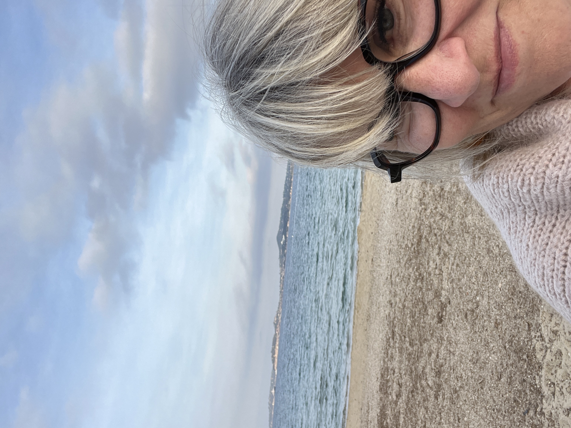 The image is an outdoor selfie taken at Grimaud, France, with the beach and sea in the background. A woman with gray hair and glasses is positioned in the right upper portion of the frame. The backdrop includes a beach, the sea, a distant shoreline, and a cloudy sky.