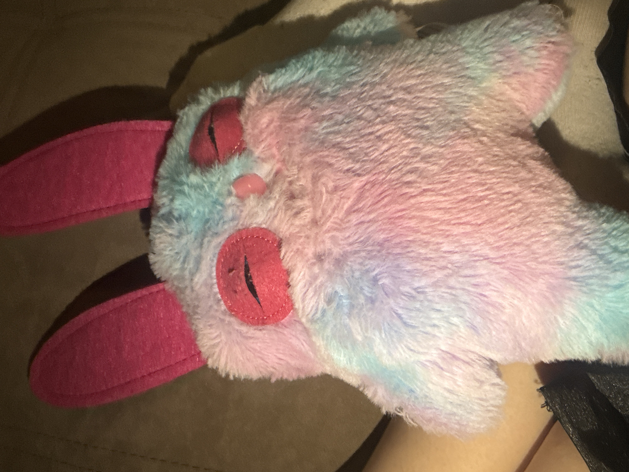 A plush, multi-colored bunny-like toy lies on a soft, brown surface, possibly a couch or bed. The toy's fur is a gradient of blue, pink, and white. It has large, pink ears made of a flat, felt-like material, and its eyes are red with black, stitched pupils. A small, pink nose is visible. The toy's pose suggests it is lying down. Part of a person's leg, with what looks like a black garment on it, is visible near the right edge of the photo. There is no indication of a location in Saint Petersburg, Russia, or any other city.