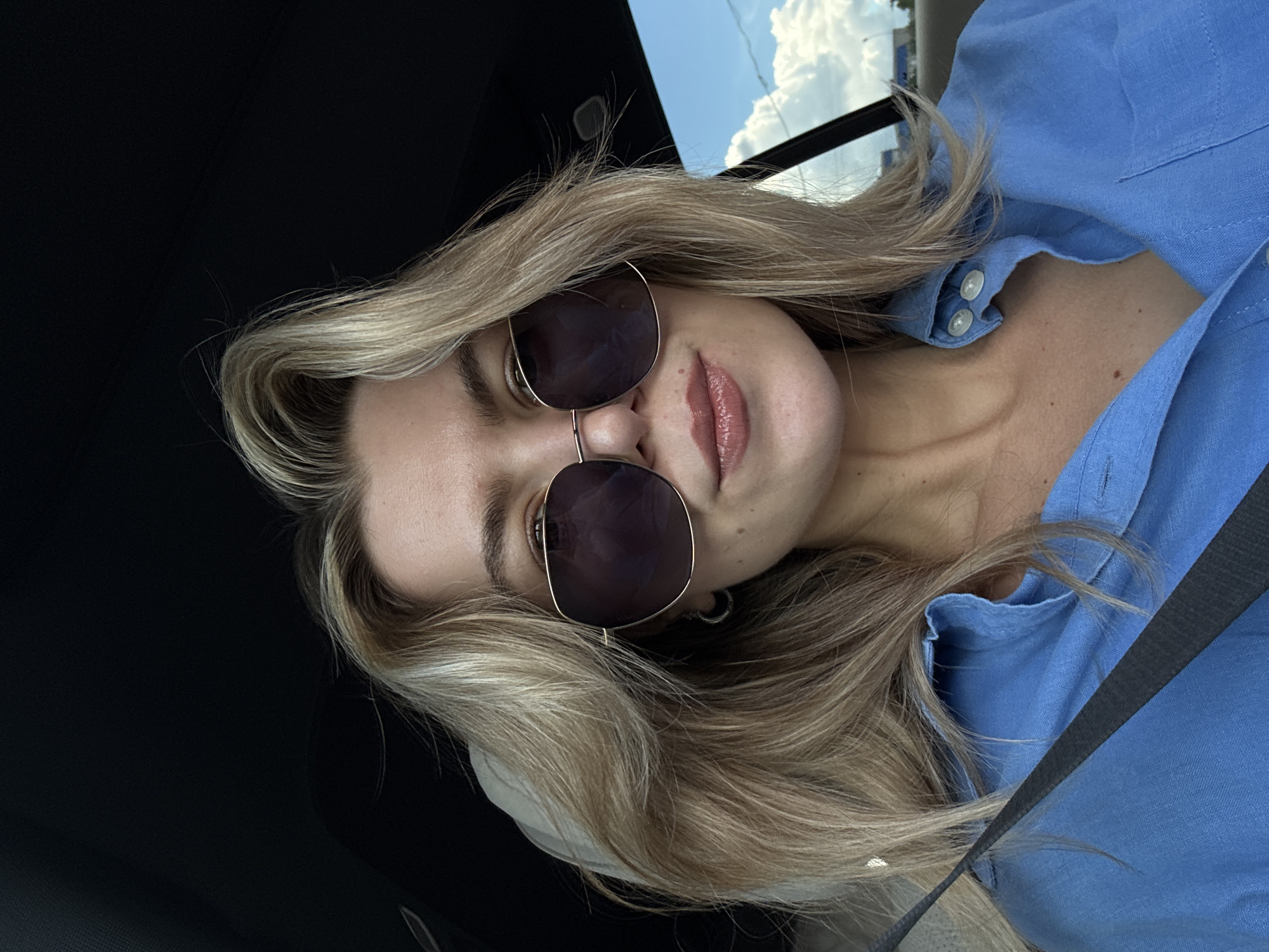 A woman with blonde hair, wearing sunglasses and a blue shirt, is positioned in a car, indicated by the black interior and the presence of a seatbelt across her body. The sky is visible through the car window with clouds present, suggesting an outdoor daytime setting. The city is not discernible from the image.