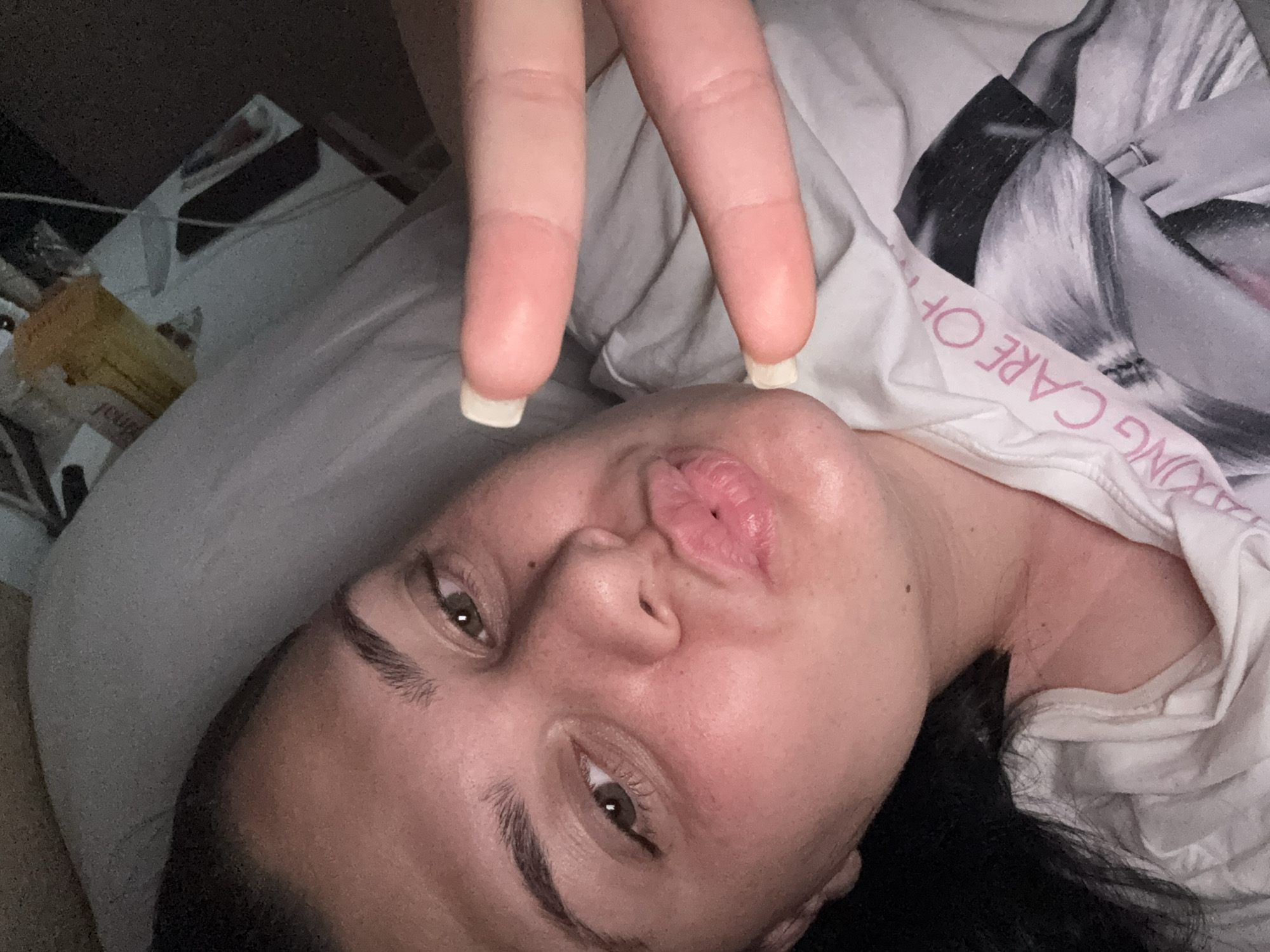 A close-up, angled selfie taken in Rogosh, Bulgaria, shows one person, likely female, lying down indoors. The individual is making a peace sign with two fingers extended towards the camera, while their lips are pursed in a playful expression, possibly a "kiss" or "duck face." Their dark hair is visible, along with brown eyes and well-groomed eyebrows. The fingernails on the visible fingers have white tips.
The person is wearing a white t-shirt that features a black and white graphic of a stylized female figure. Part of the text on the shirt is visible in pink, reading "ING CARE OF" (upside down and reversed), suggesting it is part of a longer phrase such as "TAKING CARE OF." A ring is visible on the hand within the t-shirt's graphic.
The background is dimly lit and somewhat blurry, suggesting evening or night. A grey pillow or bedding is prominent. To the left, a partially obscured yellow rectangular object is discernible. The overall mood conveyed by the pose and expression is casual and playful.