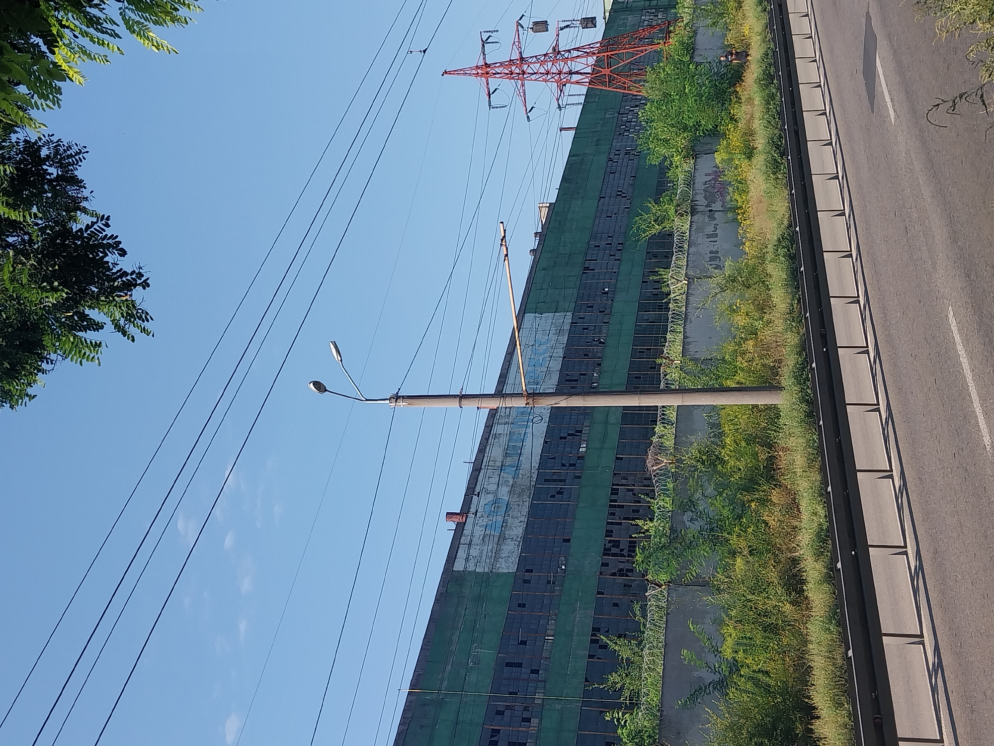 In Dnipro, Ukraine, a long, derelict industrial building with a faded green and blue facade stretches across the horizon. Its numerous windows are broken, reflecting the clear blue sky. A tall red power pylon stands to the right of the building, supporting thick electrical wires that crisscross the frame. In the foreground, a road with a metal guardrail runs parallel to the building, bordered by overgrown vegetation and barbed wire fences. A lone figure can be seen standing in the brush near the building.