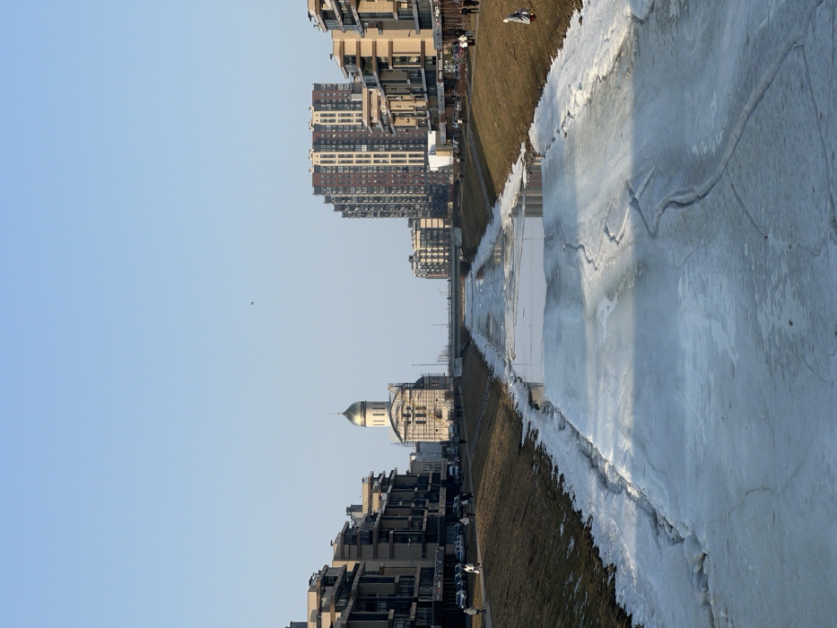 The image displays a wide-angle view of a partially frozen river, identified as the Smolenka River, on Vasilievsky Island in Saint Petersburg, Russia, under a clear blue sky. The river occupies the central foreground and mid-ground, covered by fractured sheets of white and light blue ice, with intermittent sections of dark open water reflecting the sky. Brown and sparsely green grassy slopes rise from both riverbanks, leading to paved pedestrian paths.
On the left bank, a series of modern, multi-story residential buildings with beige facades and balconies are visible. A road runs parallel to the river, featuring parked cars and several pedestrians walking on the path. In the middle distance, a concrete bridge spans the river. Beyond this bridge and on the left bank, the white-walled Cathedral of Saint Catherine stands prominently, crowned with a golden dome. Scaffolding is present around the lower levels of the cathedral, indicating ongoing construction or renovation.
On the right bank, a similar arrangement of modern residential buildings, some featuring dark red paneling, lines a paved area. Multiple individuals are scattered across both the paved paths and the grassy slopes on either side of the river. The scene suggests a late winter or early spring day, with shadows indicating clear sunlight. A small dark bird is visible in the distant sky.