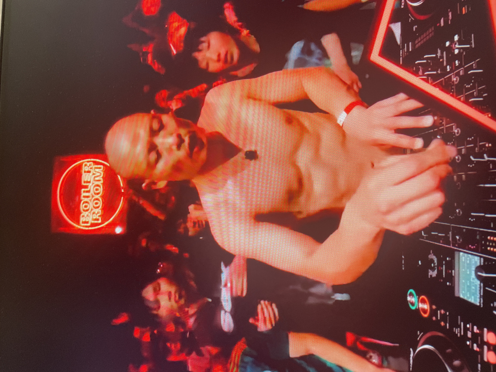 The image shows a man with a shaved head, shirtless, with his arms outstretched over a DJ console. He is wearing a red watch on his left wrist. The DJ console is black with illuminated buttons and controls. In the background, a crowd of people is visible, with some holding up their phones. A red neon sign with white lettering that says "BOILER ROOM" is displayed above the DJ. The environment appears to be a nightclub or a concert venue, with low lighting and red hues dominating the scene. The location is described as Unknown, Russia.