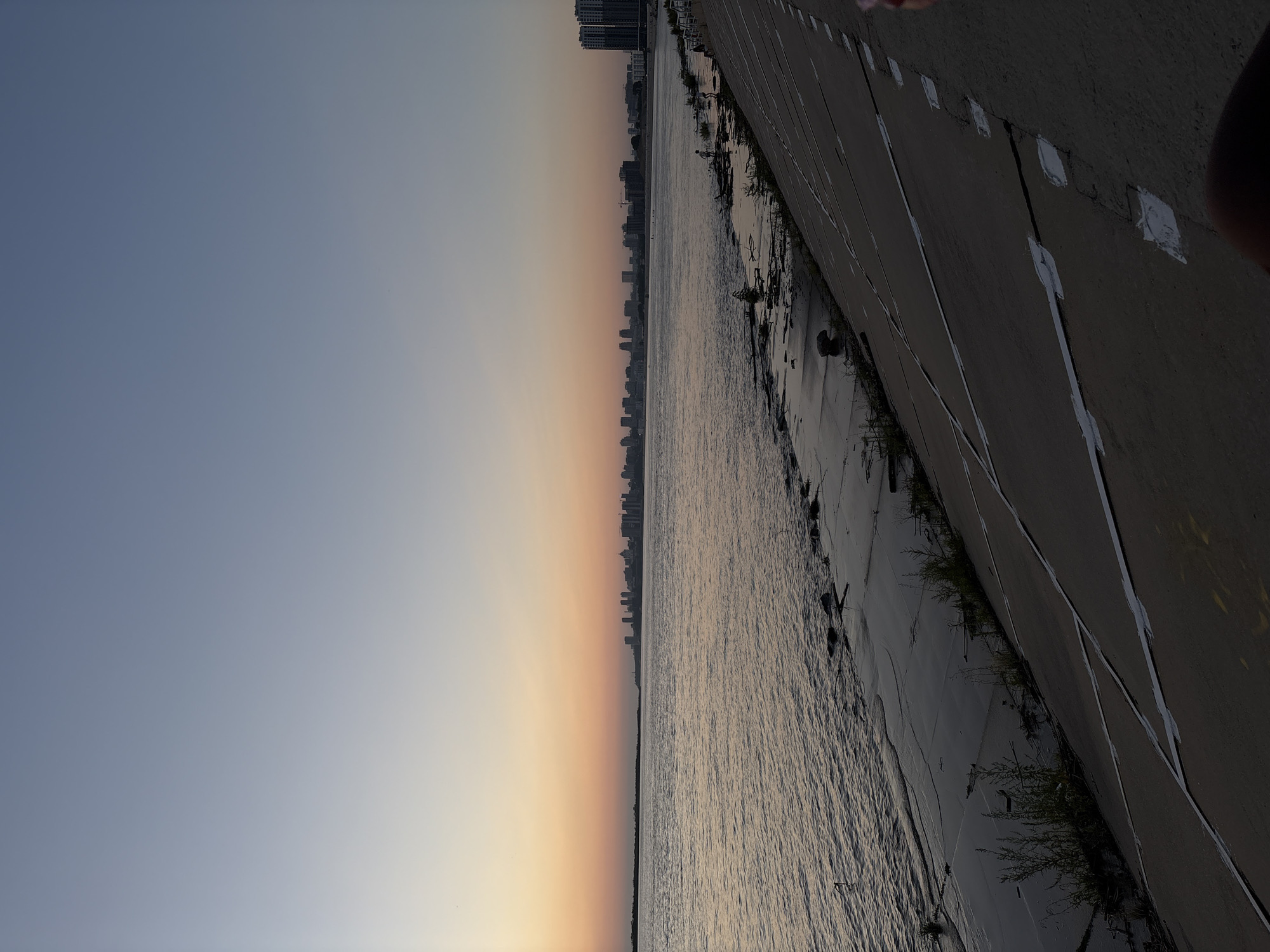 The image depicts the Amur River embankment in Khabarovsk, Russia, during twilight. The sky exhibits a gradient of soft blues and oranges as the sun sets. On the far bank of the river, the Khabarovsk skyline is visible, characterized by a cluster of multi-story buildings and skyscrapers. The river itself is wide and calm, with gentle ripples on its surface. The embankment in the foreground is a concrete structure, sloped and reinforced with white tape or markings at intervals. Sparse vegetation grows along the edge of the water. Several individuals can be observed on the embankment, some appearing to be children, engaged in activities near the water's edge. The overall scene suggests a peaceful evening by the river.