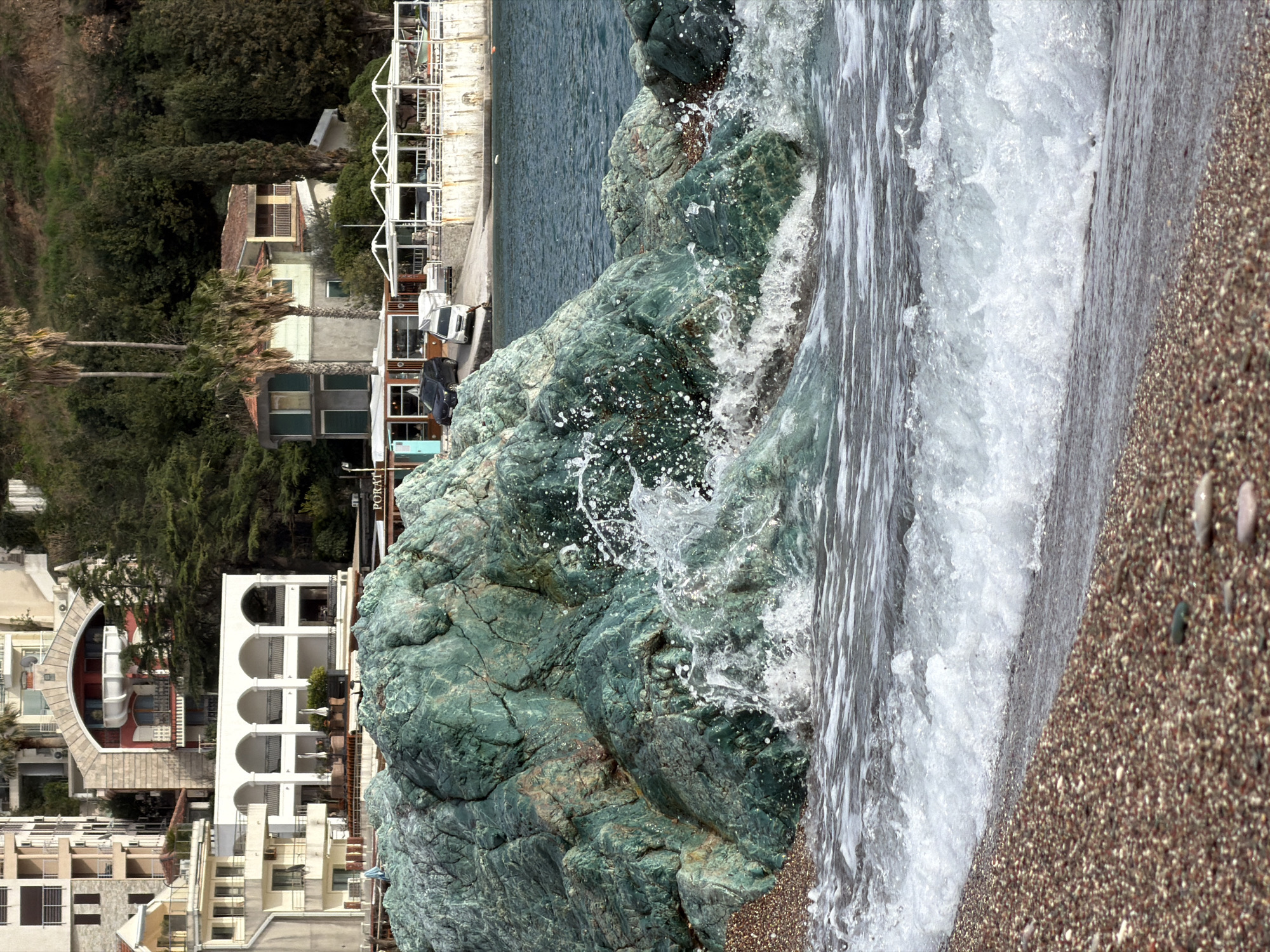 The image displays a coastal scene, rotated 90 degrees clockwise from its natural orientation, showing a shoreline with waves, large rocks, and buildings nestled among trees.
In the foreground, occupying the right side of the rotated image, is a pebbly beach with light brown, grey, and some darker colored small stones. The texture of the pebbles is clearly visible, suggesting a close-up perspective of the shoreline.
Moving left from the pebbly beach, the midground is dominated by the sea and large, dark green rocks. Waves are actively crashing against these rocks, creating a dynamic spray of white foam and water droplets that are frozen in motion. The water itself appears a deep blue-green, transitioning to white where the waves break. The rocks have a textured, somewhat weathered appearance, with variations in color from a deep teal to lighter green and hints of brown.
Further left, in the background, a series of buildings and lush green vegetation line the coast. Many of the buildings are light-colored, featuring white walls, balconies, and terracotta-colored roofs. Some structures have distinctive architectural elements, such as arches and curved balconies. Palm trees and other dense green foliage are interspersed among the buildings, climbing up a hillside. A road or promenade runs along the waterfront in front of some of the buildings, with parked vehicles visible, including what appears to be a white van and a dark car. White metal structures, possibly awnings or scaffolding for outdoor seating, are also present along this promenade.
There is some visible text on one of the buildings in the background. Part of a sign can be read as "PORAT".
The lighting suggests it is daytime, possibly morning or late afternoon, given the clear view and the quality of light. The scene appears vibrant and well-lit, indicating fair weather conditions. The overall impression is one of a picturesque coastal town or resort area.