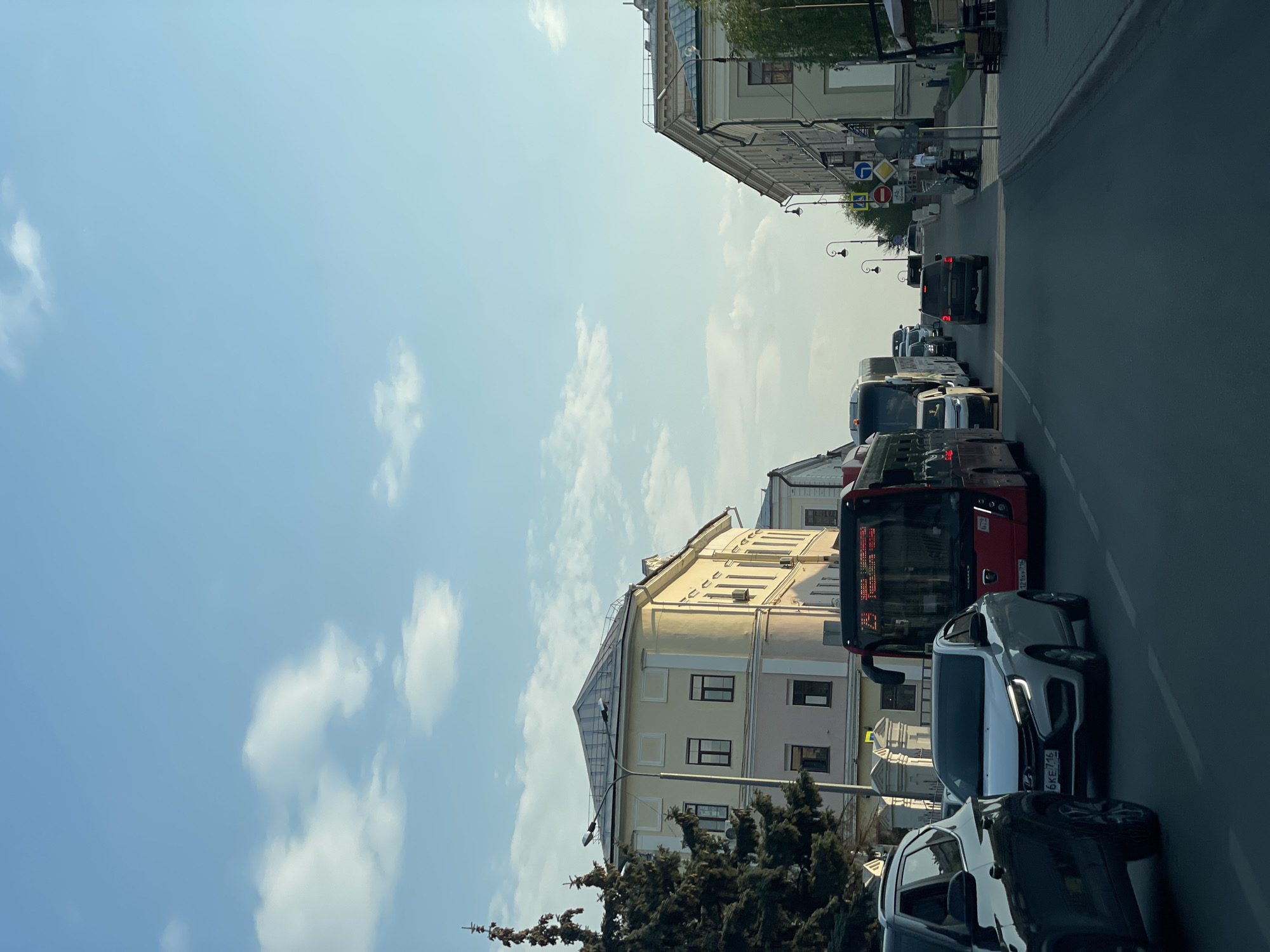 The image depicts a street scene in Kazan, Russia. The view is from street level, looking down a street with a line of stopped or slow-moving vehicles. A red bus and several cars are visible. Buildings line both sides of the street, with a mix of architectural styles. The sky is blue with scattered white clouds. Street signs and lampposts are visible along the sidewalk. At the far end of the street, a pedestrian can be seen walking on the sidewalk.