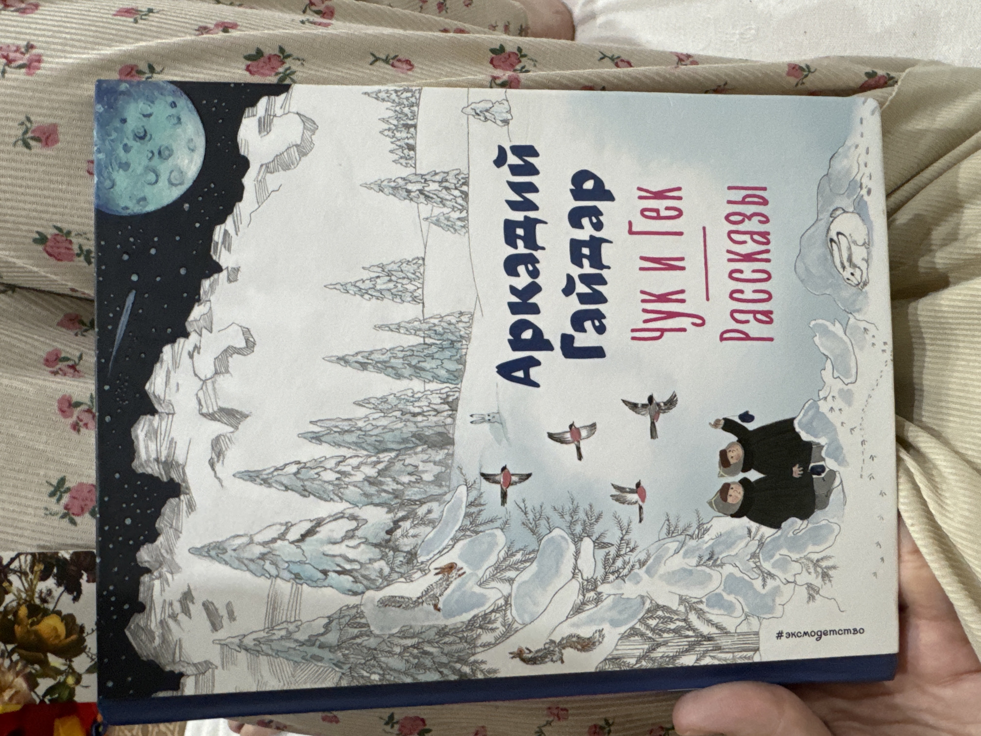 The image shows a book lying on a surface with a floral pattern. The book cover features a winter scene with snow-covered trees and a moon in the upper left corner. Several birds are depicted in the air, and two figures are seen near the snowy ground. The title of the book, written in Russian, is "Аркадий Гайдар Чук и Гек Рассказы," which translates to "Arkady Gaidar Chuk and Gek Stories." The hashtag "#эксмодетство" is visible at the bottom right corner of the book cover. There is no information about Chaikovsky, Russia present in the image.