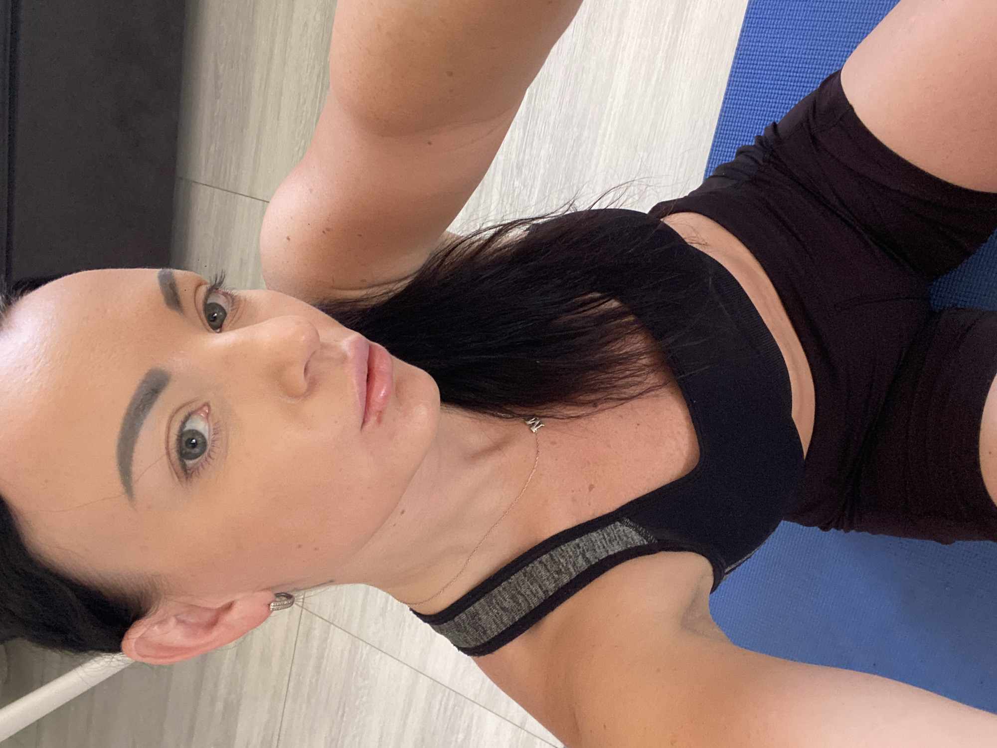 A woman is taking a selfie while on a blue yoga mat indoors. She is wearing a black sports bra and black shorts. Her hair is dark and pulled back. She has light-colored eyes and is wearing a small necklace with a letter 'M' charm. Her makeup is natural. The background appears to be a room with light-colored flooring. The scene is well-lit. There is no indication of the location being Sharm El Sheikh, Egypt.