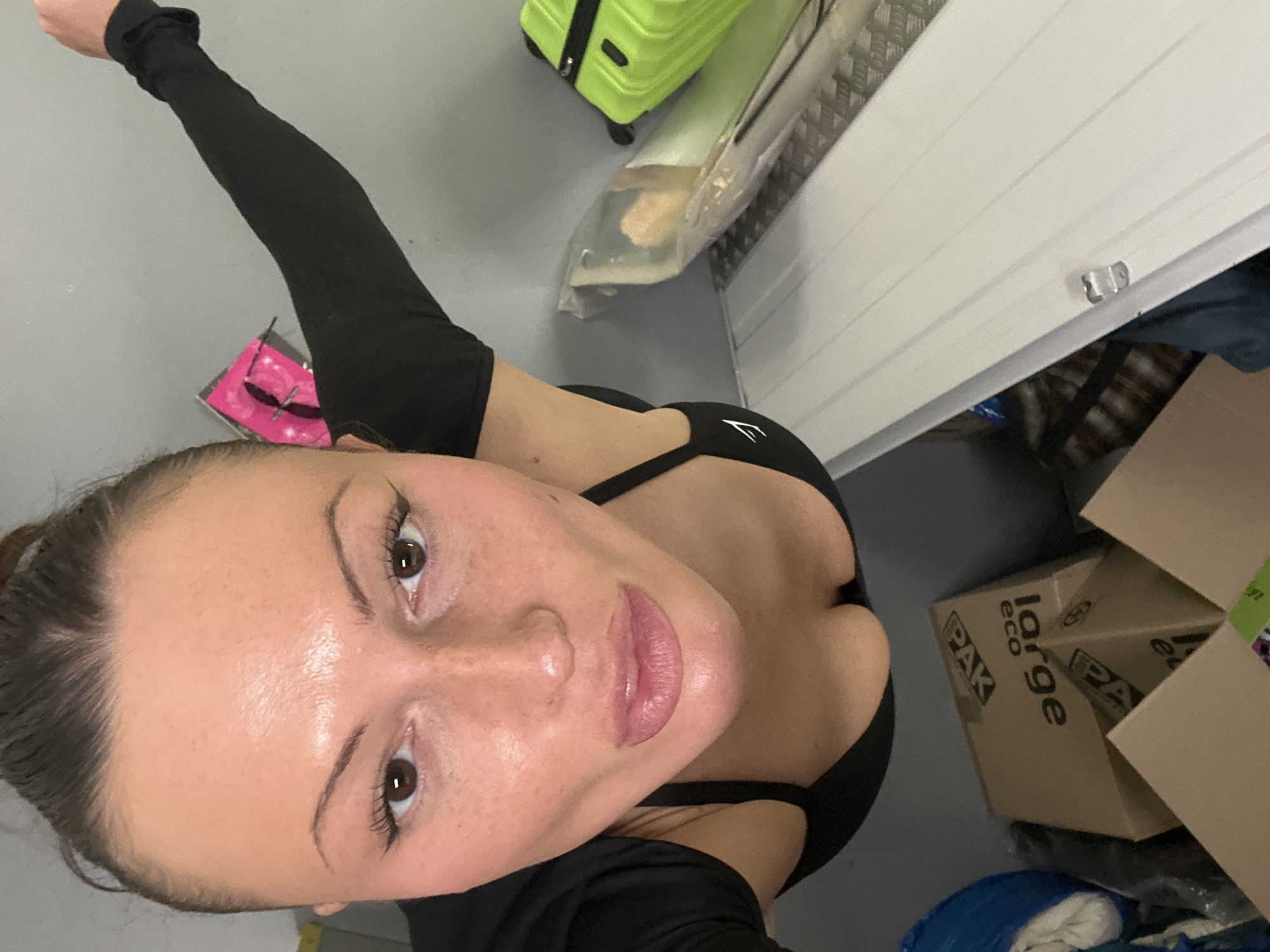 A selfie taken from a high angle shows a light-skinned woman with brown hair and brown eyes, wearing a black athletic top. She's in a room with a grey floor and a white door. Next to the door are cardboard boxes labeled "PAK eco large." In the background, there's a lime green suitcase. There's no clear indication of the location being in Tunbridge Wells, United Kingdom, based solely on the visual information available.