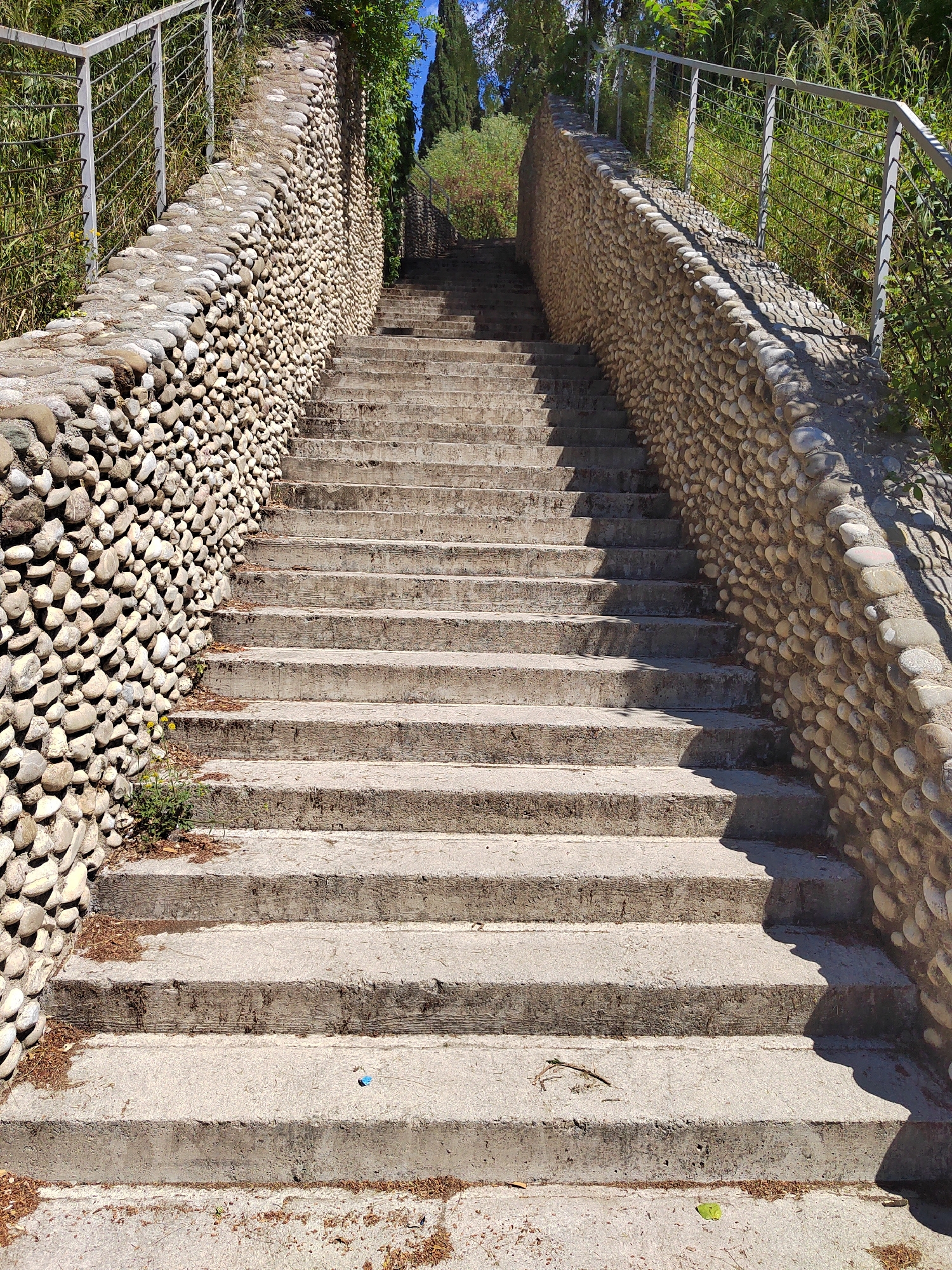 A long, straight set of concrete stairs ascends between two walls constructed of small, rounded stones. A metal railing runs along the top of each wall. Vegetation is visible beyond the walls, including trees and tall grasses. The stairs are partially shaded. The image was taken outdoors during daylight. The location is likely Podgorica, Montenegro, based on image characteristics and context.