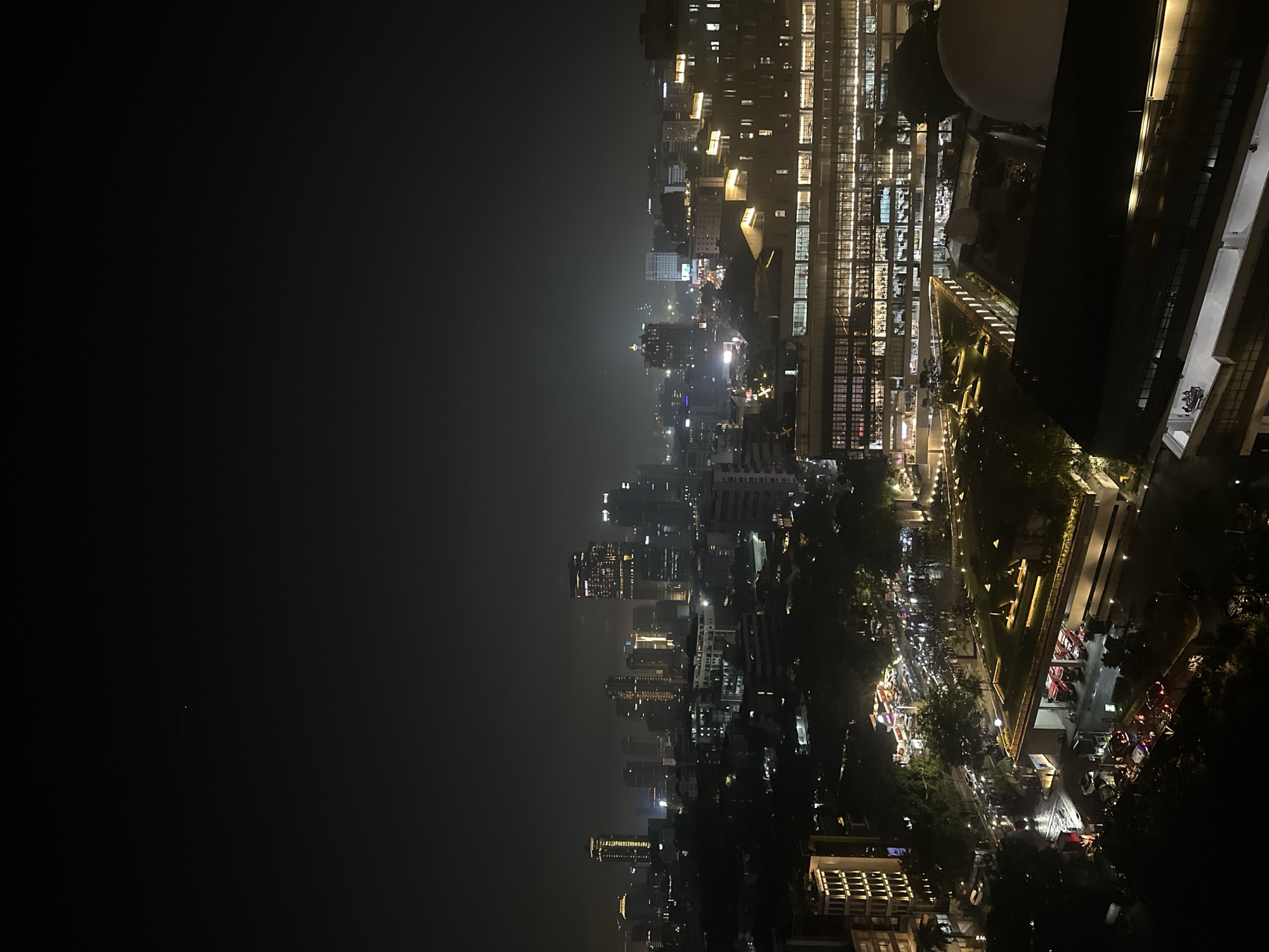 The image shows a nighttime view of Central Jakarta, Indonesia, from an elevated position. The right side of the frame features brightly illuminated buildings and streets, with a mix of modern high-rises and commercial structures. Pedestrian activity is visible along well-lit walkways and roads, along with light streams from moving vehicles. The left side of the image presents a cityscape shrouded in darkness and mist, obscuring details of distant buildings. City lights create a faint glow, indicating the presence of a dense urban environment.