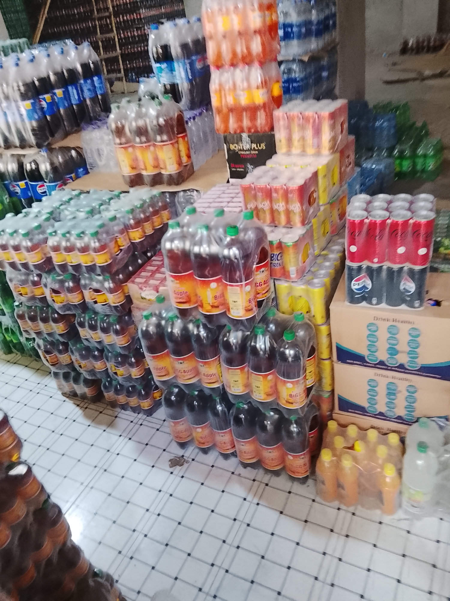 The scene depicts a storage area or small wholesale outlet, potentially within the Gulgasht Colony in Pakistan. The location is densely packed with stacks of beverages, including bottled soda (Pepsi), fruit juices (apple, peach), energy drinks (Booster Plus), and bottled water. Stacks of canned beverages are also visible. Several cardboard boxes labeled "Drink Healing" are present near the cans. The beverages are arranged on a checkered floor. The background contains additional, densely packed stock, possibly within a larger shelving unit.