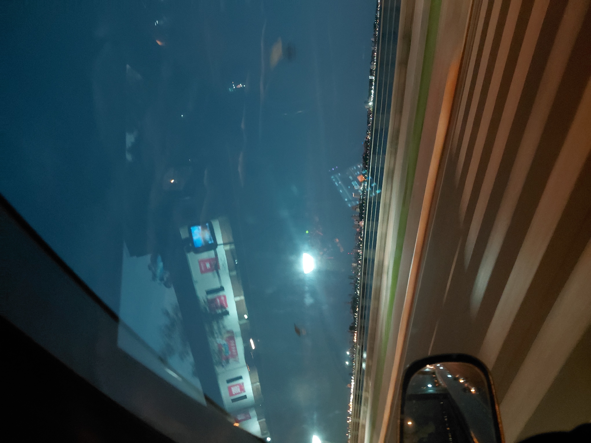 The image depicts a view from inside a vehicle, looking forward through the windshield and to the side through the side mirror. The time of day appears to be dusk or night, with a dark blue sky overhead.
In the foreground, the road is visible, marked with white stripes for a pedestrian crossing, indicating the vehicle is either stopped or moving slowly. The side mirror on the left side of the frame reflects lights from the road and surroundings, suggesting the vehicle is in motion.
In the mid-ground, a body of water, likely a river or bay, stretches horizontally across the frame. Beyond the water, a city skyline is visible, illuminated by numerous lights. The city appears to be Encarnación, Paraguay, given the context.
Reflected in the windshield, there is an image of a building with signage, possibly advertising or informational displays. The reflection is somewhat distorted, typical of viewing through a vehicle window. The building appears to have red and white elements. There are also some blurred lights and shapes in the reflection, including what looks like a traffic light or a screen with blue and white elements.
The overall impression is of a city at night, seen from a moving vehicle, likely crossing a bridge or driving along a waterfront road.