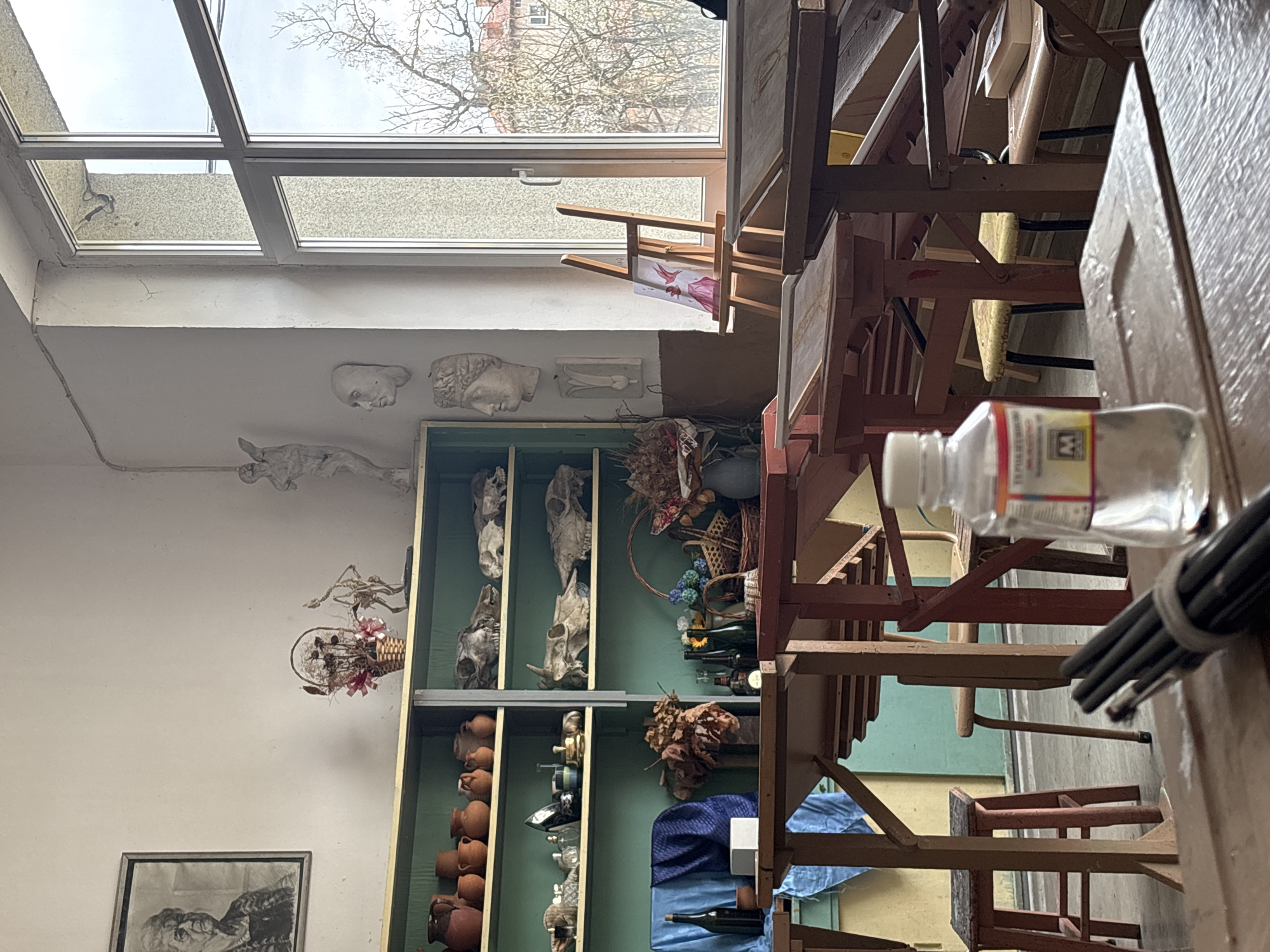 The image shows the interior of what appears to be an artist's studio or atelier, possibly in Troyan, Bulgaria, given the mention of the city in the prompt. A large multi-paned window at the top of the frame allows daylight to enter. Below the window, a white wall is decorated with plaster casts of faces and limbs, as well as a framed drawing or painting. A tall, green-painted cabinet is filled with various objects, including animal skulls, pottery vessels, bottles, and decorative items like dried flowers and baskets. The foreground is dominated by rows of wooden easels and chairs, arranged in a tight formation. A clear plastic bottle and a bundle of paintbrushes are situated near the front. A blue cloth is draped over an object near the bottom of the cabinet. The scene evokes a sense of organized clutter, typical of a working studio environment.