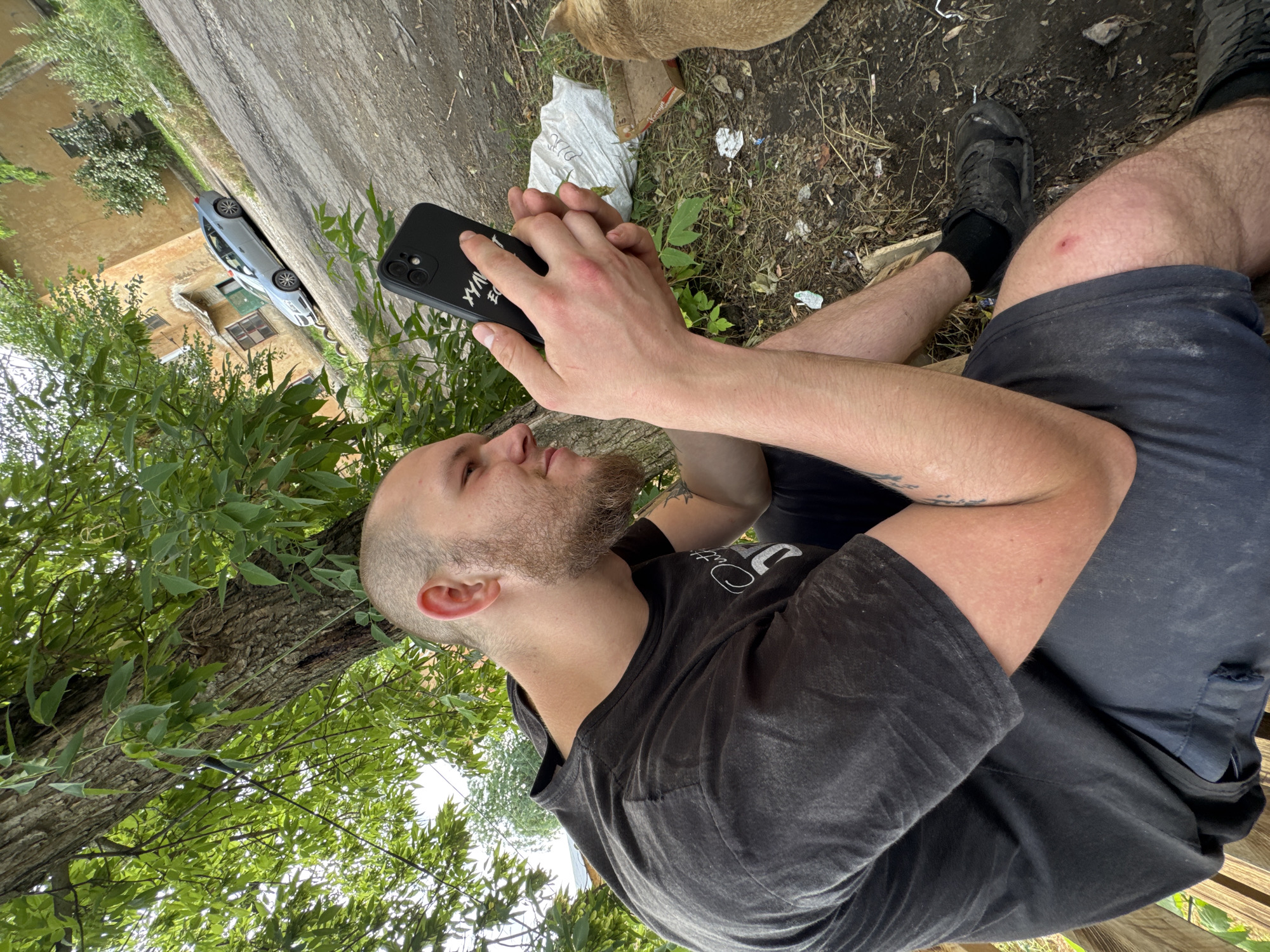 A man with a shaved head and beard sits outdoors, looking at his phone. He is wearing a black t-shirt and dark shorts. His phone is in his hands, and he appears to be interacting with it. Behind him, a tree with green leaves provides shade. In the background, there is a street, a light-colored building, and a car. The ground is dirt and littered with some debris, including a white plastic bag and a cardboard box. A dog, partially visible, is to the right of the man.