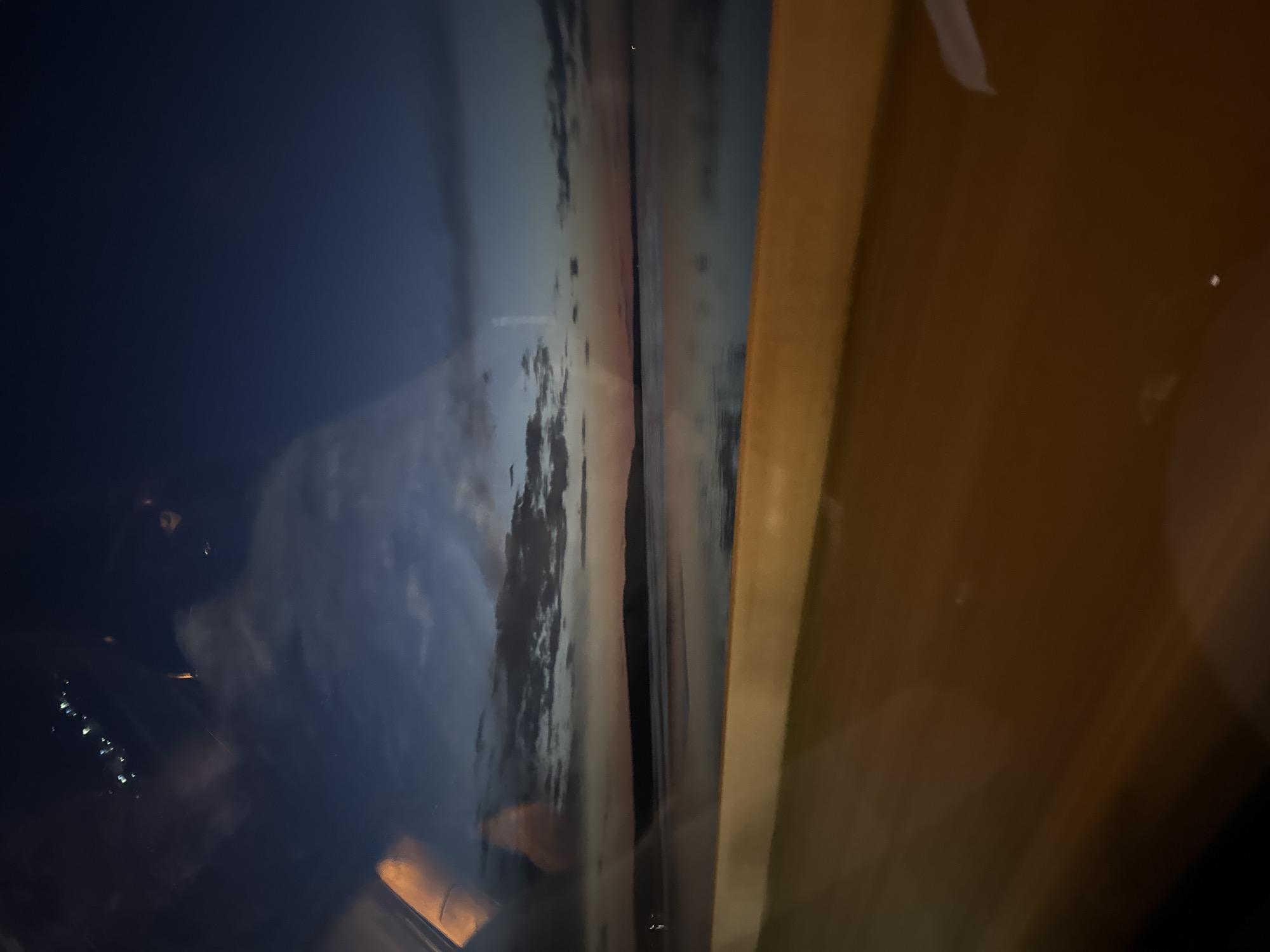 The image appears to show a reflection in a window or glass surface. The reflection depicts an outdoor scene with a sky that transitions from a dark blue to a lighter, cloudier area, potentially at sunset or sunrise. A portion of a building or structure is visible on the right, appearing brown or beige, and possibly made of wood. The dark area on the left side might be a city at night, with scattered lights. There is no definitive proof that this location is in Chaikovsky, Russia, as the image alone does not provide concrete geographical markers.