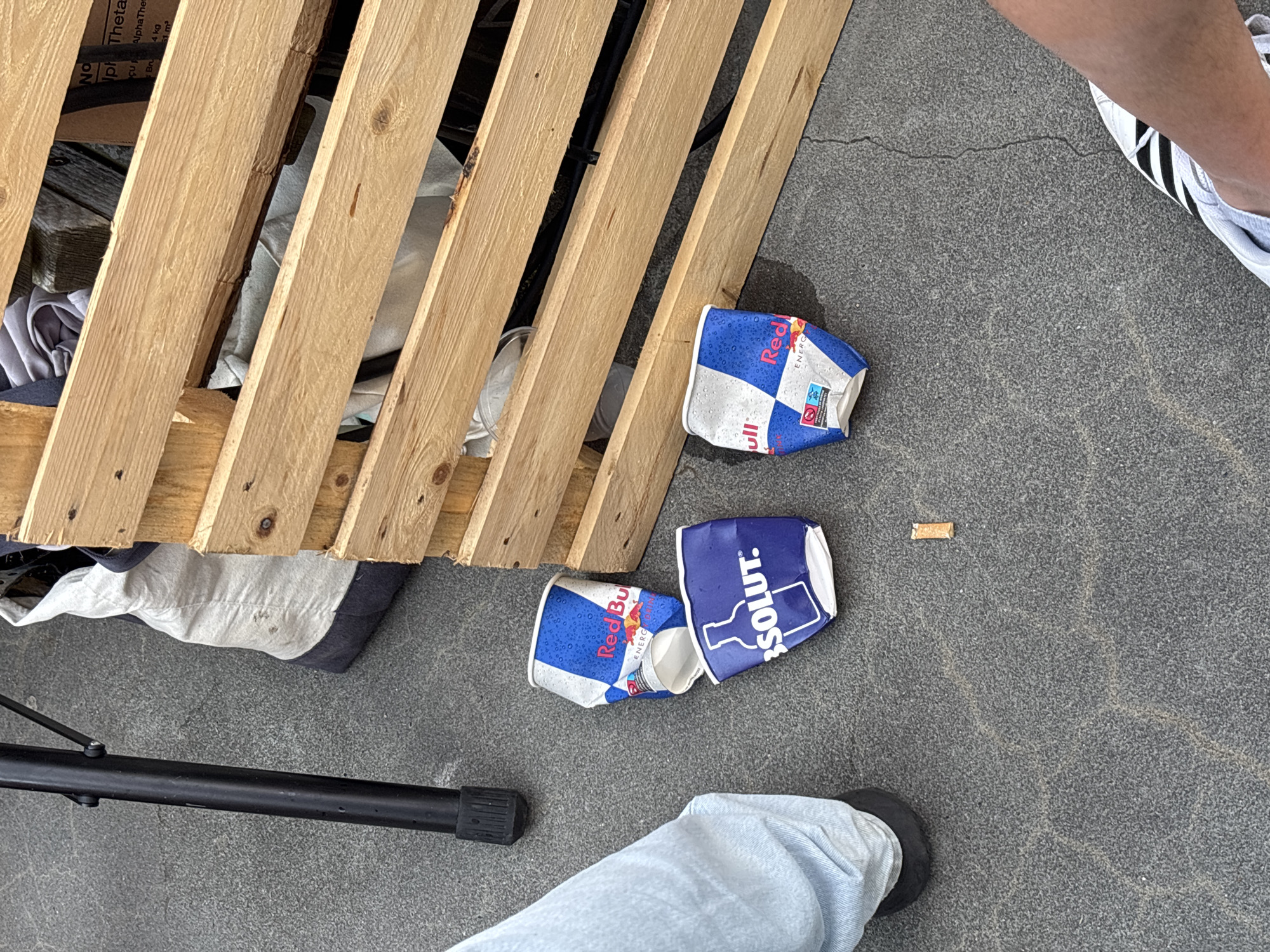 An outdoor scene on a paved surface, likely Sundbyerne, Denmark, features three crumpled beverage cups and a cigarette butt. Two of the cups are Red Bull branded, with their characteristic blue and red design. The third cup is branded with Absolut, a clear spirit. The cups are scattered on the ground. To the left, a person's legs, clad in light blue jeans and black shoes, are visible, suggesting they are either standing or sitting. A black metal tripod leg is also visible in the left portion of the frame. To the right and behind the cups, a wooden pallet is positioned, and some fabric items, possibly clothing or bags, are visible within the pallet's structure. The pavement exhibits cracks, indicating wear and tear. In the bottom right corner, another person's legs, wearing white athletic shoes with black stripes, are visible. The lighting suggests it is daytime.