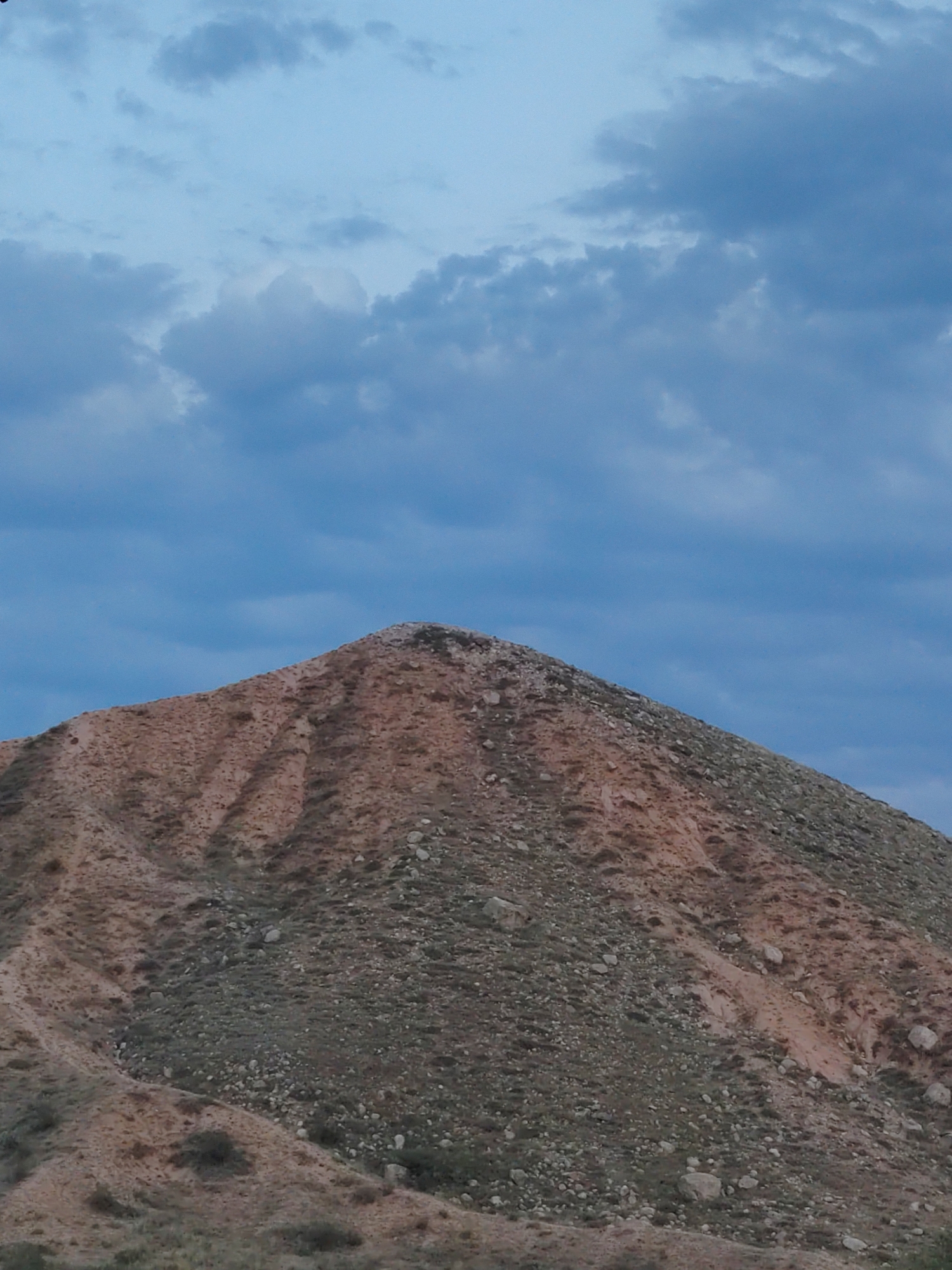 The image depicts a mountain peak with a rocky, arid slope covered in sparse vegetation. The sky above is overcast with blue and grey clouds. The scene appears to be a natural landscape. There are no people or specific urban features visible in the image, so it cannot be definitively located in Cholpon-Ata, Kyrgyzstan, or any other specific urban location. The description is limited to the visible elements in the photograph.