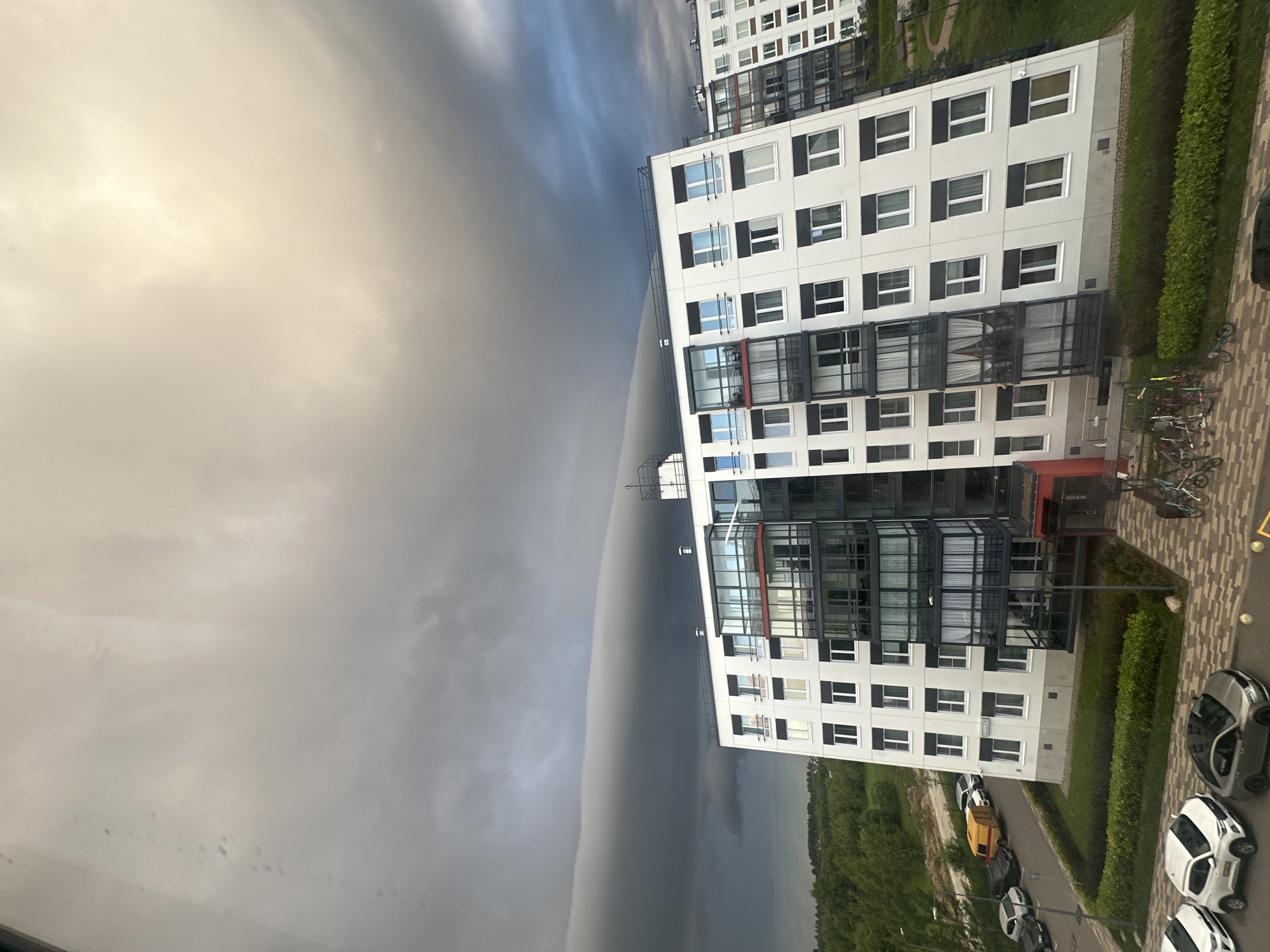 The image shows a residential building in Vsevolozhsk, Russia, under a dramatic, stratified cloud formation. The building is modern, with white facades, numerous windows, and glass-enclosed balconies. To the left of the building, there is a street with several parked cars, including a yellow utility vehicle. A patch of green trees is visible in the background. The sky is dominated by heavy, dark clouds forming a distinct horizontal band across the frame, with lighter, brighter clouds above. The overall scene suggests an approaching storm.