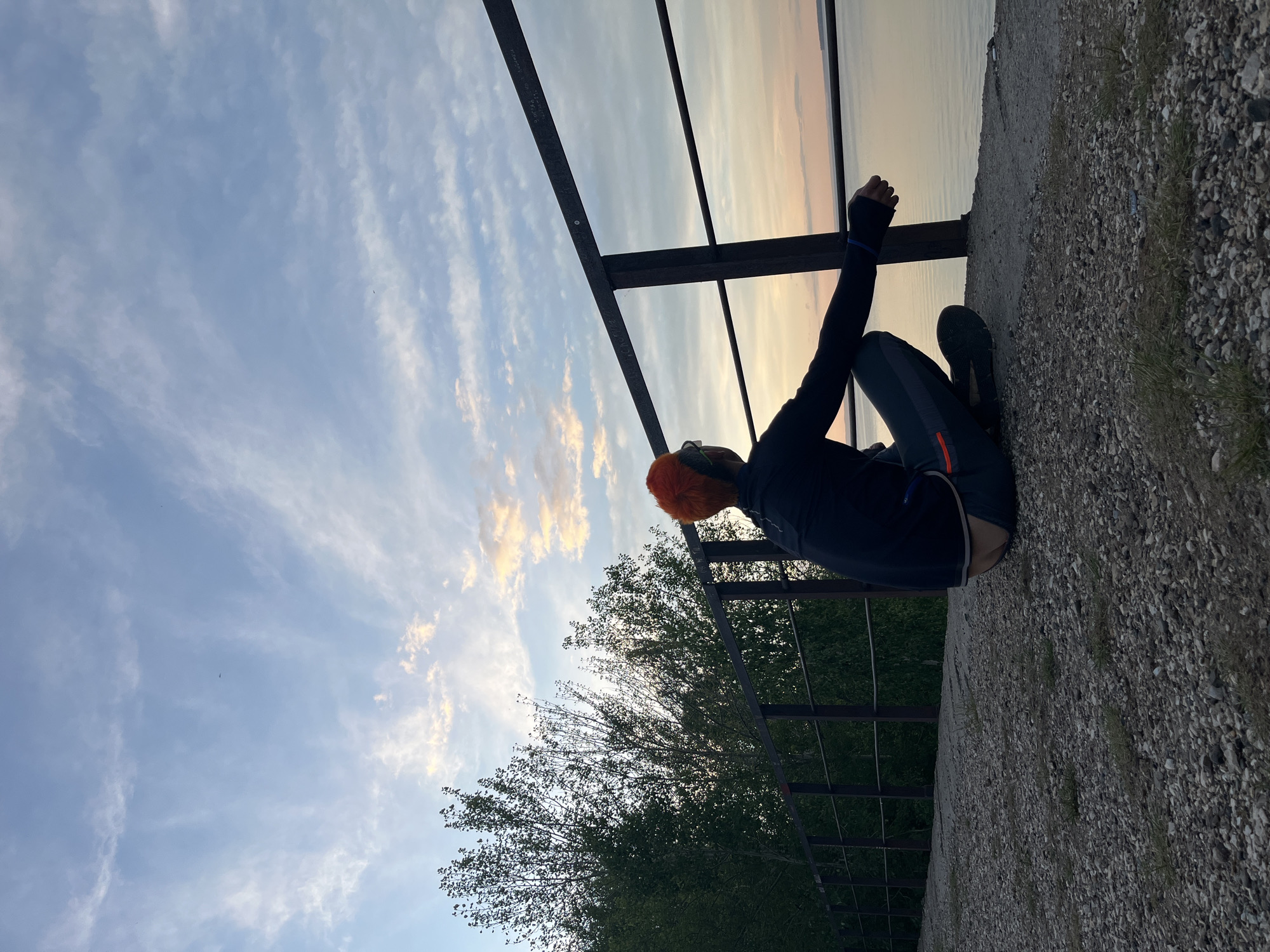 A person with orange hair sits on a concrete ledge next to a dark metal railing structure. They are wearing dark clothing with blue accents and dark shoes. The background shows a sky with light clouds and the silhouette of trees. The surface next to the ledge is covered in small rocks and grass. The location is Chaikovsky, Russia.