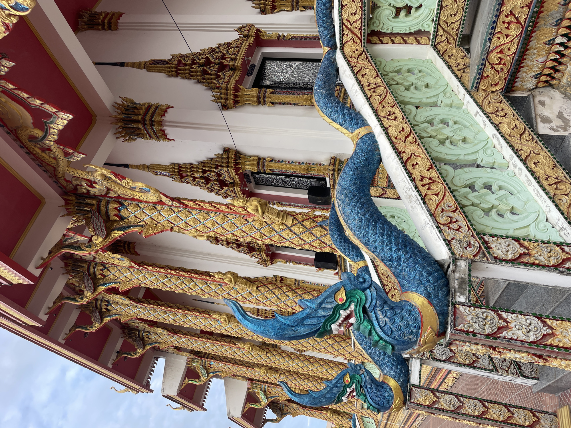 The image shows the exterior of a Buddhist temple in Pattaya City, Thailand. The temple features ornate gold and red decorations, with numerous columns adorned with intricate carvings. Two blue, dragon-like serpent sculptures with green accents are prominently displayed along the railings of a staircase. The sky is partly cloudy.