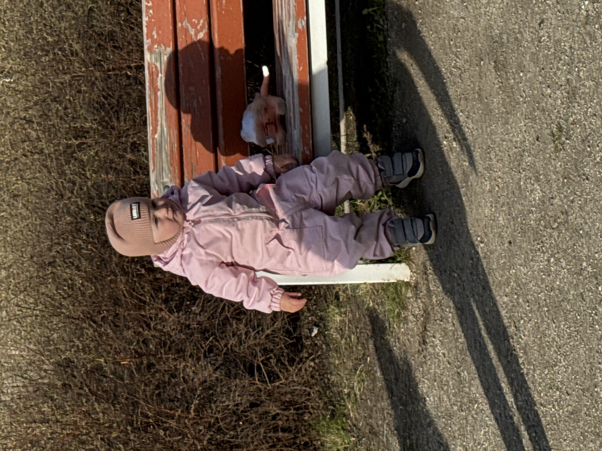 A child wearing a pink full-body suit, a light pink beanie, and dark shoes lies on a wooden park bench under bright daylight. The child's head is positioned towards the left, with their body extending across the bench. A rectangular patch on the beanie clearly displays the text "URBAN."
The bench is constructed from reddish-brown painted wood, which appears worn with areas of peeling paint, and features some white or light-colored slats. Peeking from underneath the bench's slats is a light-colored stuffed animal, possibly a cat or rabbit, distinguished by an upright tail.
The scene is an outdoor setting, likely a park or public area in Yekaterinburg, Russia. To the left of the bench, the ground is covered with dry, brown vegetation, such as grass or low bushes, indicating either early spring or late autumn. To the right of the bench, there is a paved path made of small, grey gravel or pebbles. Long, distinct shadows are cast across the pavement and partially onto the bench, suggesting strong sunlight and a clear sky, indicative of midday or early afternoon. The child's facial expression is calm.