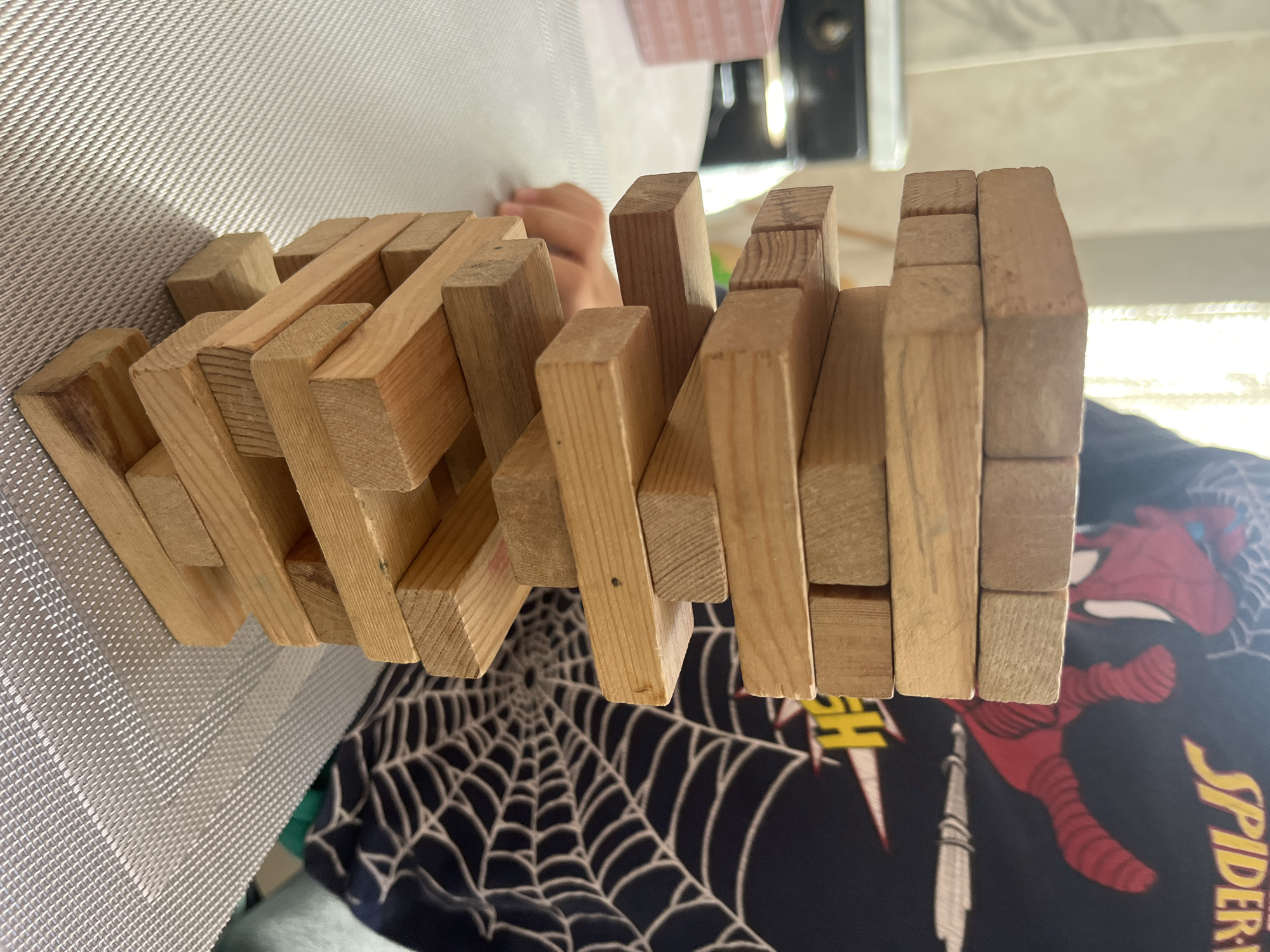 The image shows a wooden puzzle, seemingly assembled from small wooden blocks. The puzzle rests on a patterned surface, revealing a design of a spider web and a stylized depiction of Spider-Man. The presence of the spider web and Spider-Man design indicate the location is likely a bedroom, or play area of a child. There are no indicators to pinpoint the specific location to Kaspiysk, Russia.