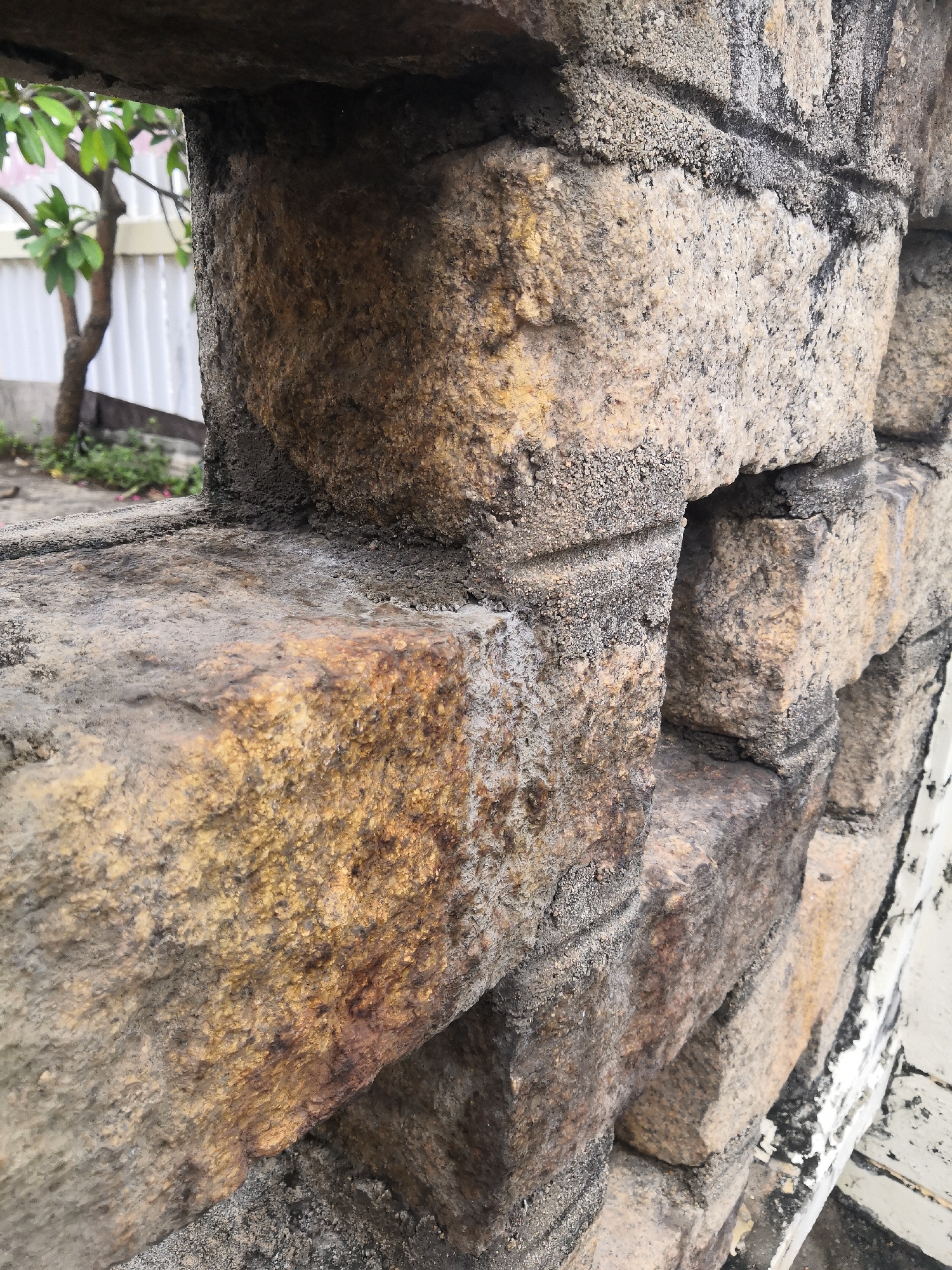 The image shows a close-up of a weathered stone wall. The stones are rectangular and stacked, with gray mortar between them. The stones have a rough texture and vary in color from light beige to dark brown. Some stones appear damp. There are small gaps or openings between some of the stones. In the background, through an opening in the wall, a tree and a white fence are visible. The location is in the city of Bac Nha Trang Ward, Vietnam.