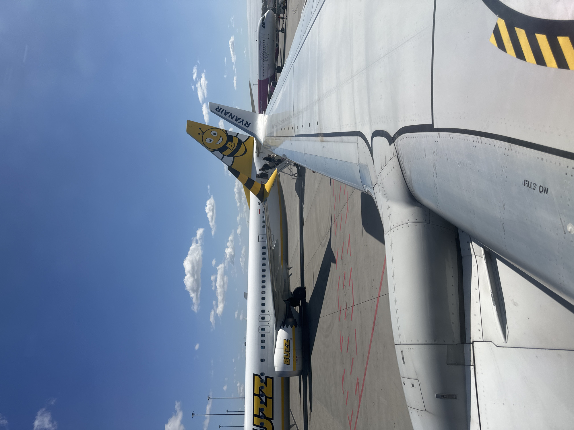 The image shows the wing of an airplane at Wrocław Airport in Wrocław, Poland. The wing belongs to a Ryanair aircraft. Another aircraft, with purple lettering, is visible in the background. The sky is blue with some clouds. A Buzz airplane is also visible. A person is seen near the Buzz plane. Markings on the ground include red painted lines. Lettering on the Ryanair wing says "FUS ON."