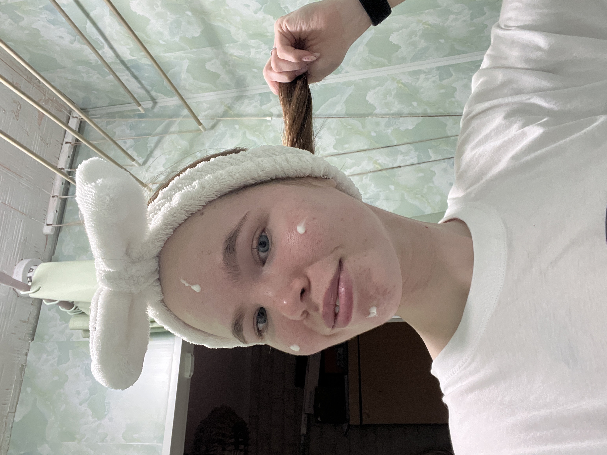 A pale-skinned woman wearing a white t-shirt and a white headband with bunny ears is visible from the chest up. She has light brown hair, some of which is being held up by a hand with a black watch. There are small dots of white cream on her face. The background appears to be a bathroom with green marble patterned wall and ceiling, with a retractable clothes dryer rack hanging from the ceiling. The location is indicated as Chaikovsky, Russia.