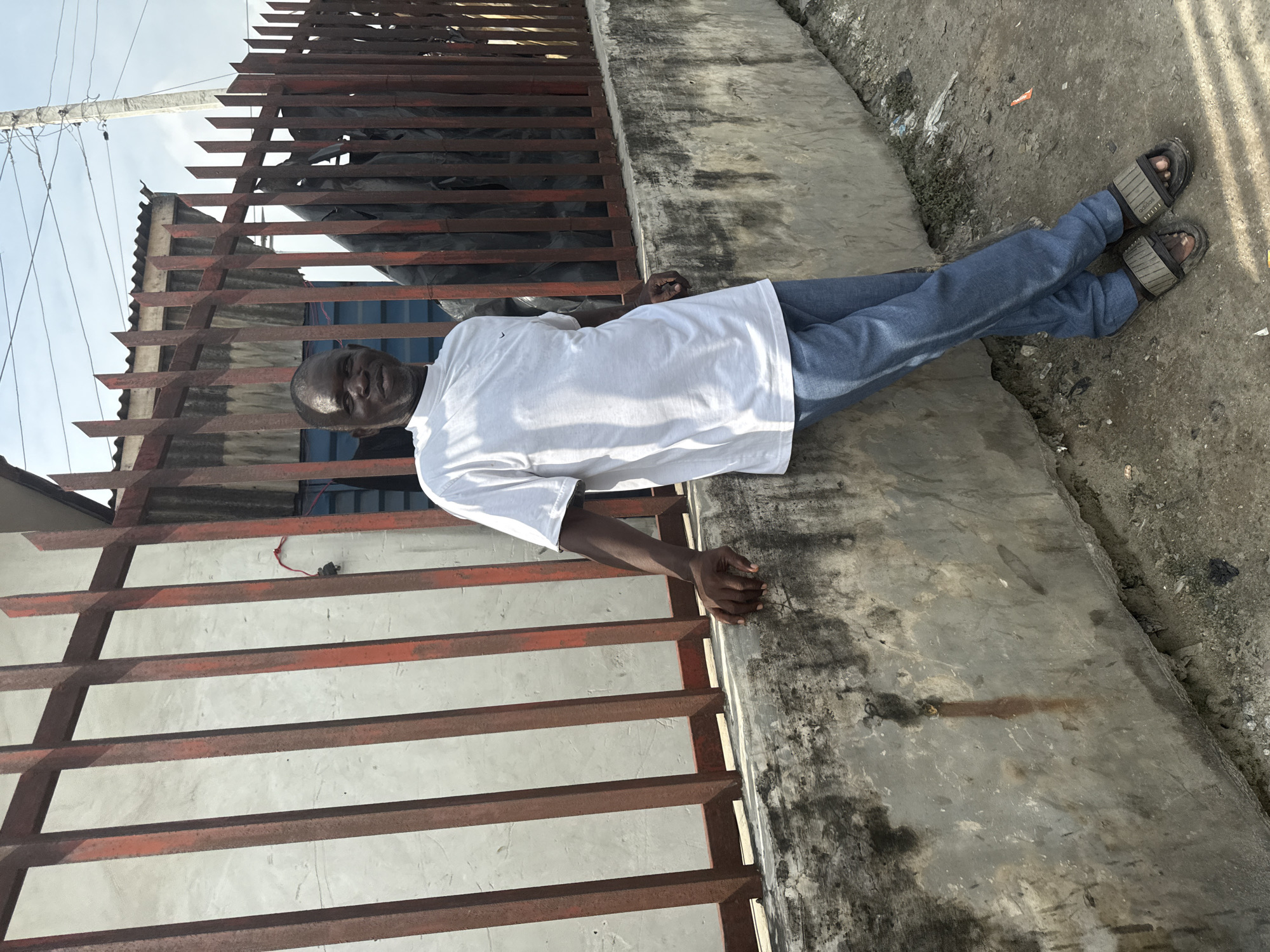 An outdoor, full-body portrait captures a dark-skinned man leaning casually against a rusty metal fence, looking directly at the viewer with a neutral expression. He appears to be middle-aged with very short hair. He is dressed in a plain white short-sleeved t-shirt, blue jeans, and dark sandals. His left hand rests on a concrete ledge or wall running parallel to the fence, while his right arm is relaxed by his side.
The scene is bathed in bright daylight, indicating clear, sunny weather. The background features the red-brown, weathered metal fence with horizontal bars, behind which is a darker area, possibly covered with a tarp, and a concrete structure with a corrugated roof. A utility pole with wires is visible on the left side of the frame, set against a bright, clear sky. The ground beneath the man's feet is a worn, dirty concrete surface, showing some cracks and debris, suggesting an outdoor, possibly urban or semi-urban environment in Ado, Nigeria. Notably, the man's sandals clearly display the brand name "Adidas." The image captures a moment of quiet observation in an everyday setting.
