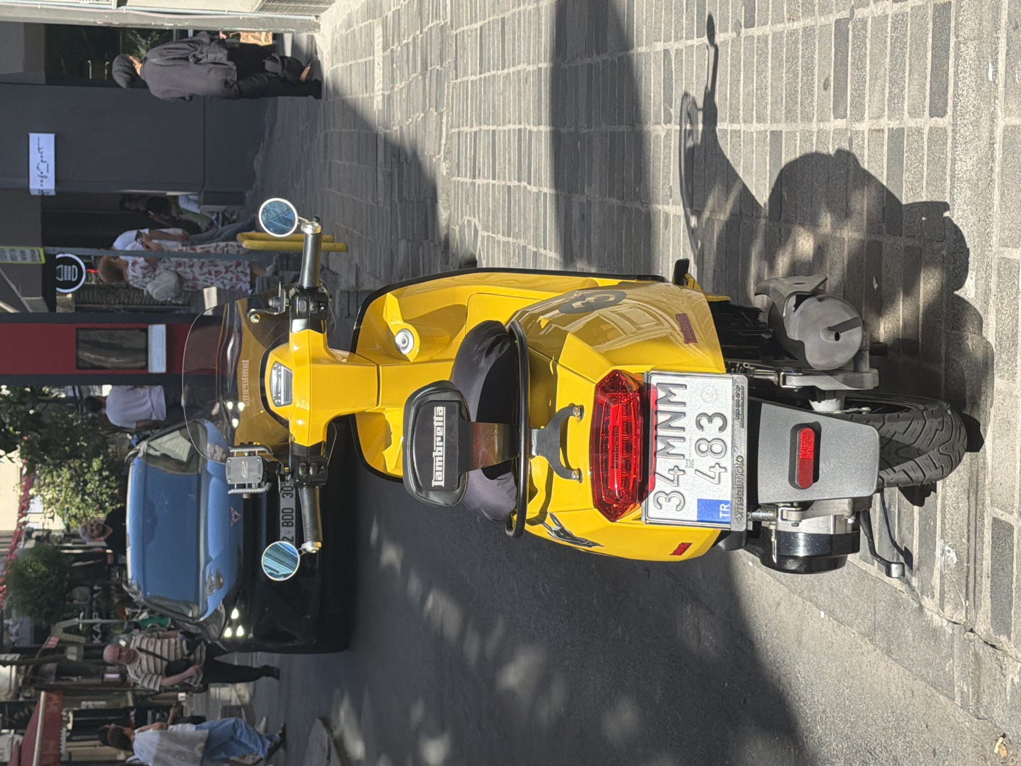 In Şişli, Turkey, a bright yellow Lambretta scooter is parked on a stone-paved street. The scooter faces away from the camera, displaying its rear, including a red taillight and a license plate that reads "34 MNM 483 TR". The scooter has a black padded backrest with "Lambretta" written on it and two round mirrors attached to its handlebars. Behind the scooter, a black SUV is visible, also facing away, with its headlights on. Several people are walking on the sidewalk to the left of the scooter and in the background, including a woman in a patterned dress and a person in a black headscarf. The street is lined with buildings, some with storefronts. Sunlight casts shadows on the pavement.