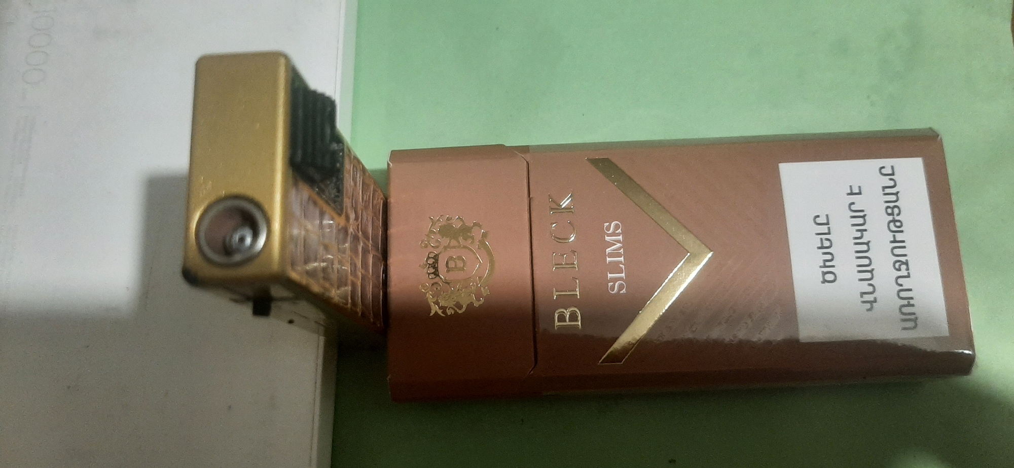 A gold lighter is placed next to a rose gold cigarette pack. The lighter has a textured pattern and a circular opening. The cigarette pack has the brand name "BLECK SLIMS" printed in gold lettering. There is also text in Armenian on a white sticker on the pack. The objects are on a light-colored surface and a green surface. The overall scene is not specific to Yessentukskaya, Russia.