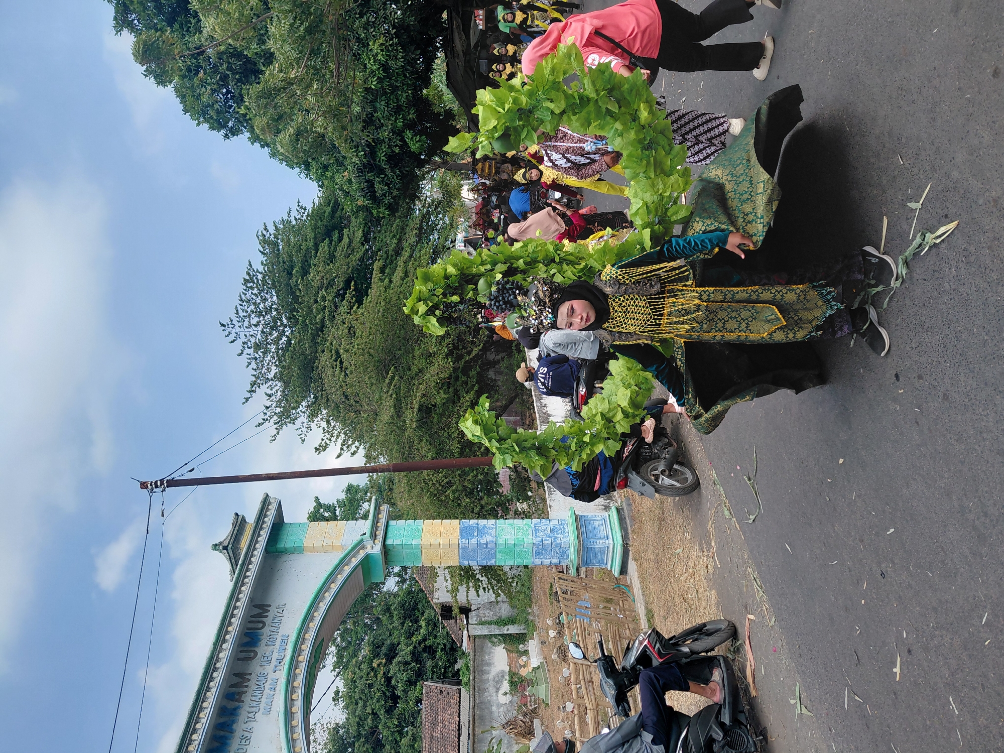 In Bimo, Indonesia, a parade is underway, possibly for a cultural celebration or festival, in front of an arched monument reading "MAKAM UMUM". The monument is decorated with colorful tiles in blue, yellow, and green, and is flanked by pillars and lush trees. In the foreground, a young person in a striking, ornate costume adorned with greenery and gold is the central focus. This costume appears to be part of a larger prop, possibly depicting a plant or a mythical creature. The individual wears a dark head covering, an elaborate headdress, and a flowing green and gold outfit. Behind them, other participants in similar festive attire, including some in yellow and patterned clothing, are visible, along with onlookers and motorcycles. The scene takes place on a paved road, with some debris and fallen leaves scattered on the surface, suggesting a street event. The sky above is partly cloudy.