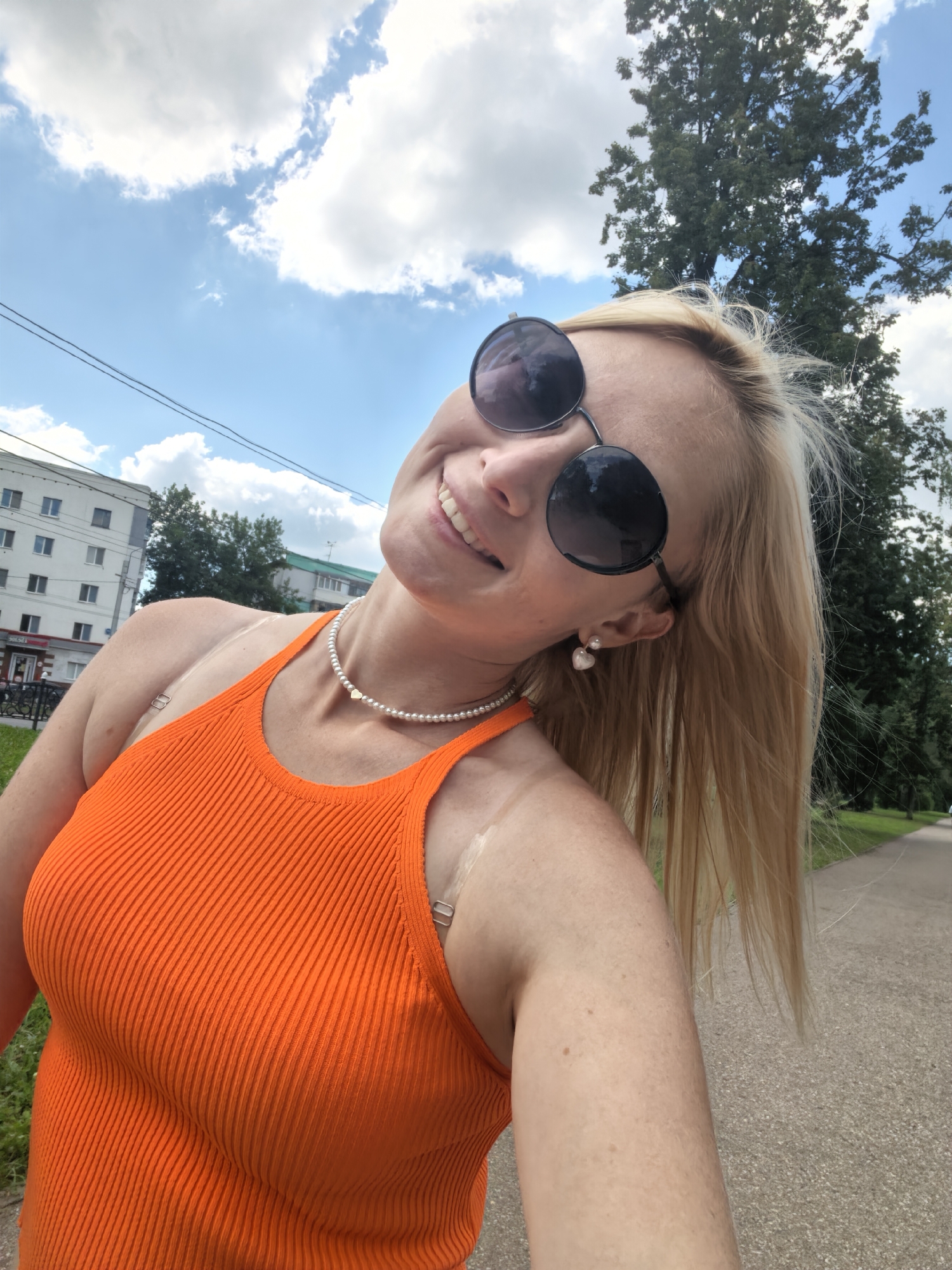 In Sterlitamak, Russia, a blonde woman with fair skin and light freckles is captured in a selfie outdoors. She is wearing a bright orange ribbed tank top and large, round, dark sunglasses. A pearl necklace with a small heart pendant adorns her neck, and she has pearl earrings shaped like hearts. Her head is tilted back, and she has a wide smile, showcasing her teeth. Her blonde hair is long and flows down her back. The background reveals a cloudy blue sky and a large tree. To the left of the frame, a light-colored apartment building with multiple windows is visible, along with some power lines. The ground appears to be a paved path with some gravel scattered on it, suggesting a park or sidewalk area. There are faint tan lines on her shoulders, indicating recent sun exposure.