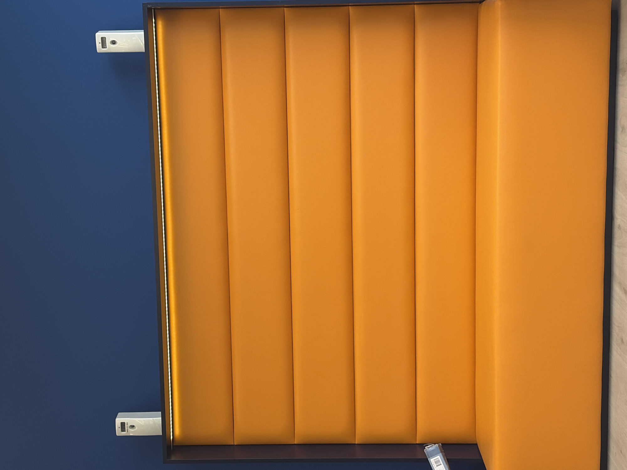 An orange upholstered bench seat is positioned against a dark blue wall. Above the bench, a strip of LED lights is integrated into a dark frame. Two white rectangular devices, possibly sensors or smart home devices, are mounted on the wall above the orange bench. The wall and bench are well-lit, highlighting the texture of the upholstery and the smooth finish of the wall. The floor beneath the bench appears to be light-colored wood. The scene suggests an indoor environment, possibly a waiting area or a modern lounge space.