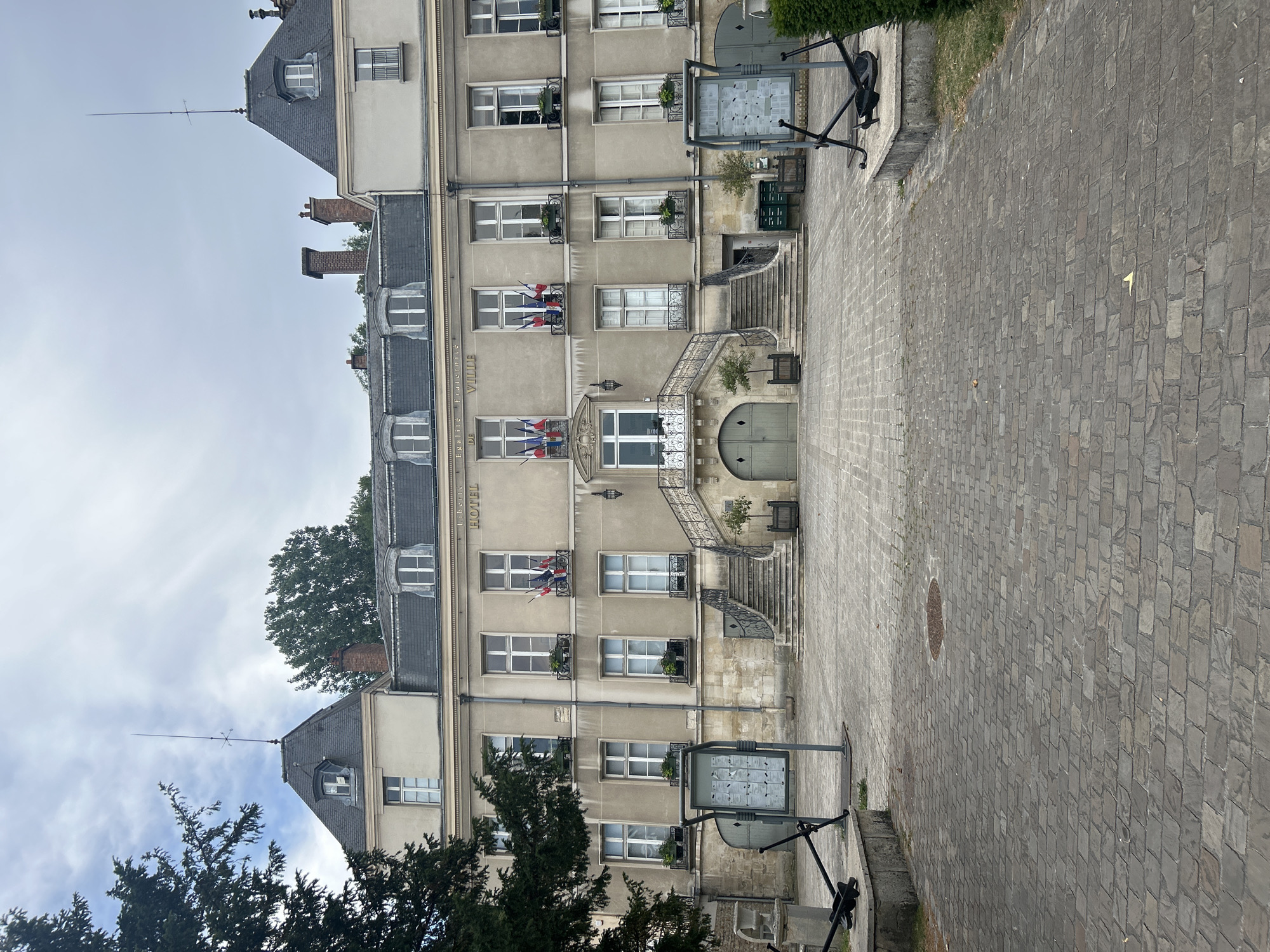 The image depicts the Hôtel de Ville (Town Hall) of Le Port-Marly, France. The building is a two-story structure with a mansard roof, featuring multiple windows on each level. French flags are displayed in some of the upper windows. A grand staircase with ornate railings leads to the central entrance on the first floor. In front of the building, there is a cobblestone courtyard with several informational kiosks and decorative anchors. Lush green trees frame the scene on the left side. The sky is overcast, suggesting a cloudy day.