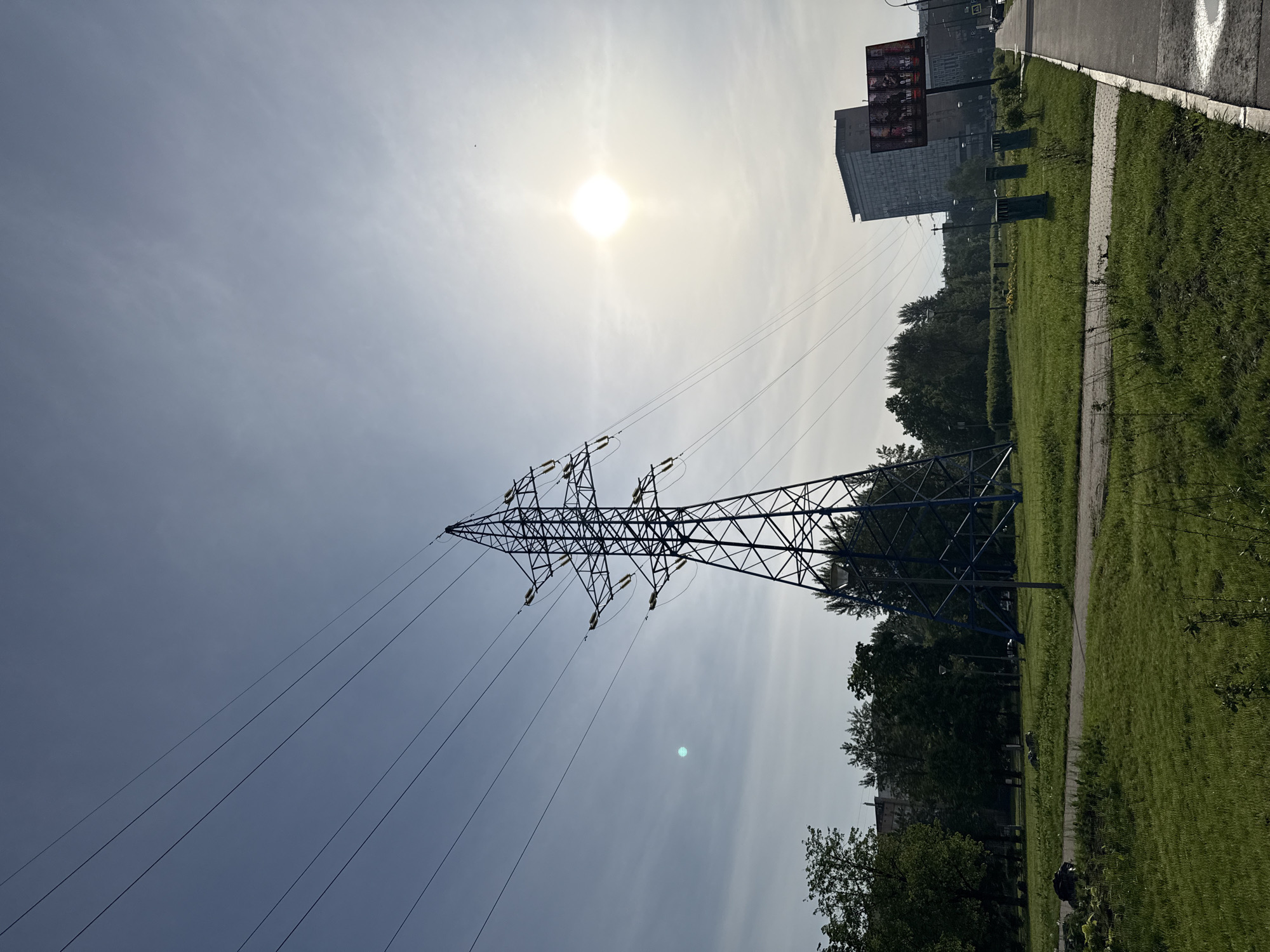 The image shows a high-voltage power transmission tower in an urban environment. The tower is a lattice structure made of dark metal. Power lines extend from the tower towards the sky. The sky is bright, with the sun visible. In the background, there is a building and greenery. The location is likely in Moscow, Russia, as per the user's request. A sidewalk and road are also visible. There is no evidence of people in the image.